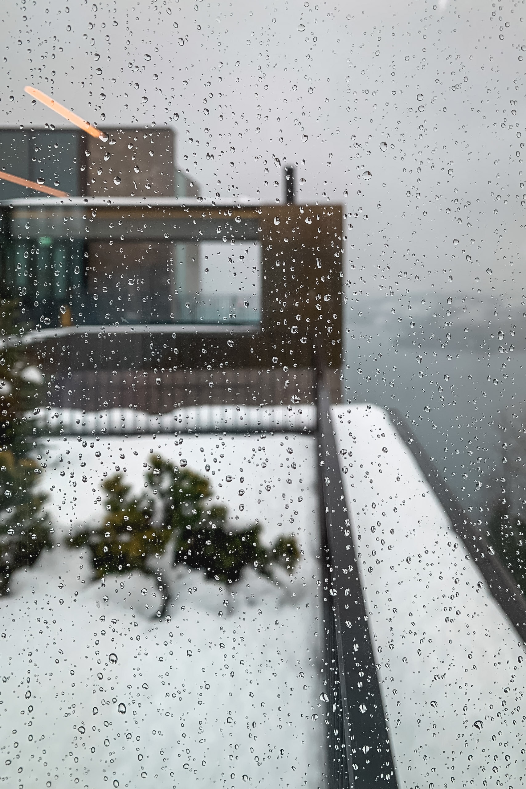 Rain-covered window reveals a snow-dusted rooftop terrace with wooden railings and evergreen plants in soft, overcast Alpine weather