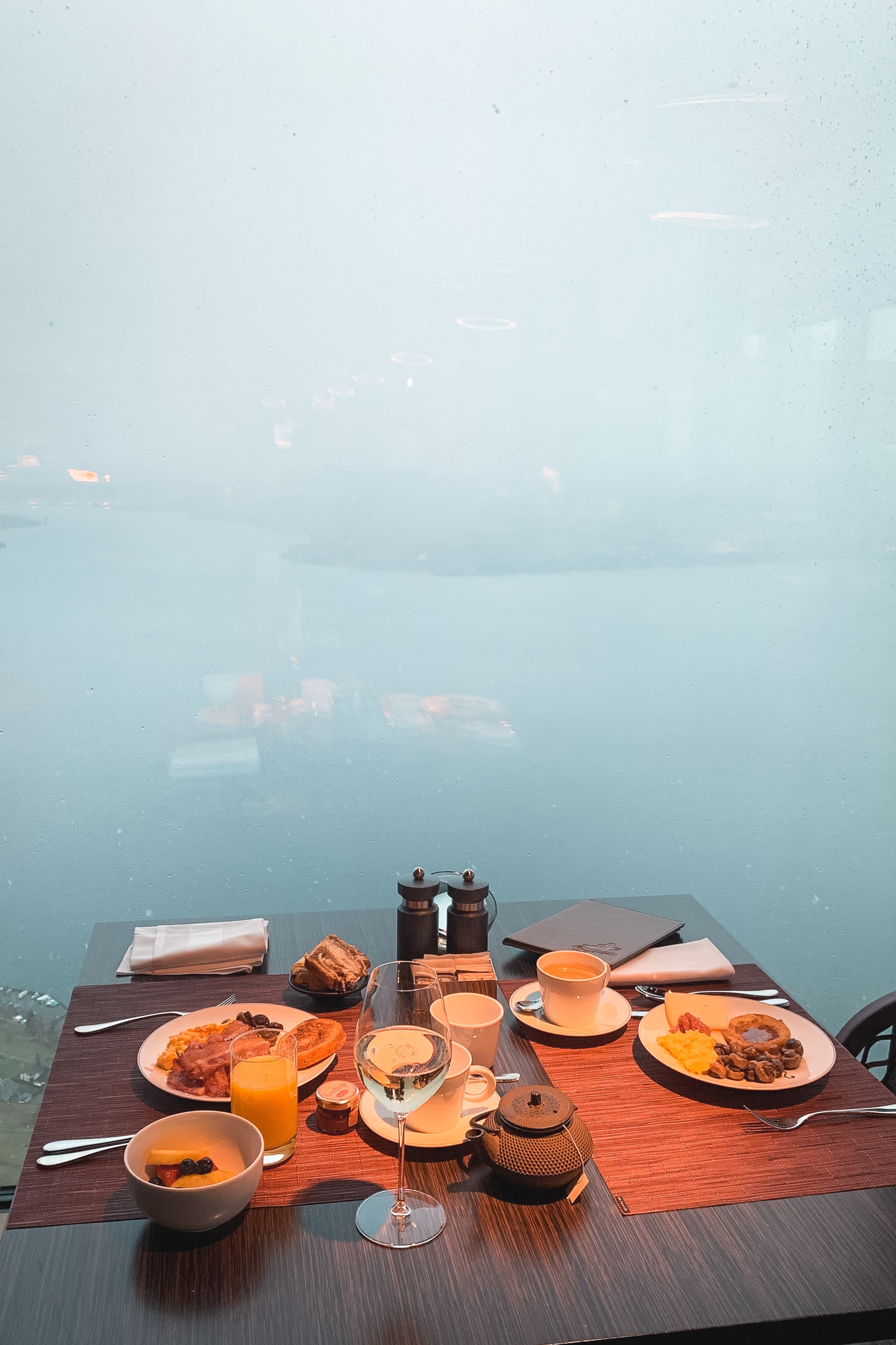 A breakfast table overlooking a misty alpine lake displays plated food, orange juice, and tea amid dense fog obscuring distant mountains and buildings