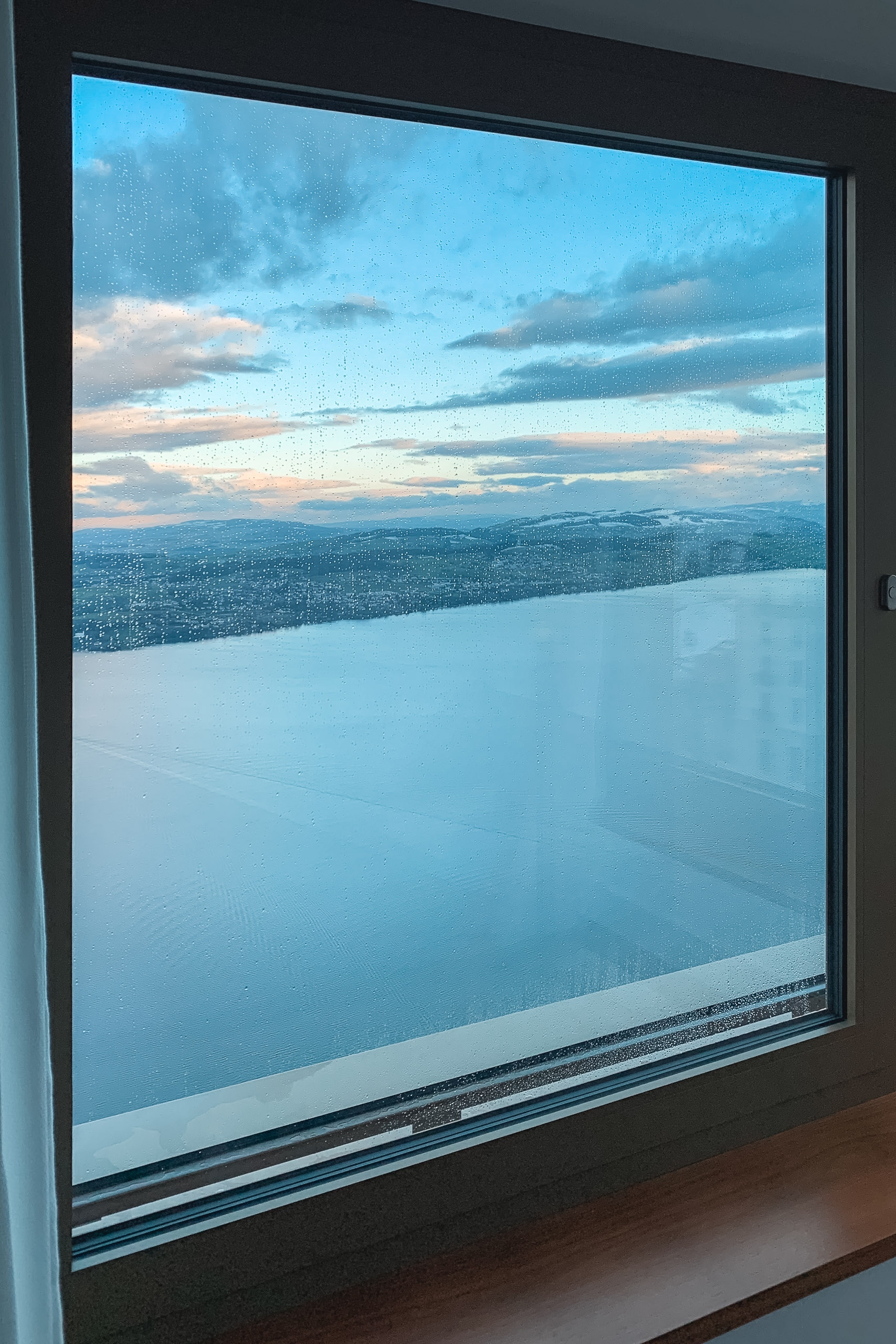 Large window framing a twilight alpine landscape with snow-covered peaks, forested valleys, and a calm lake reflecting soft blue-gray evening light