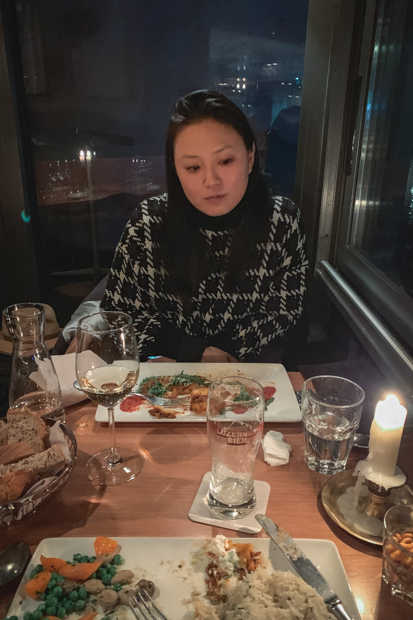 A woman in a patterned black and white sweater sits at a candlelit dining table beside windows overlooking a nighttime city skyline, with plated food and wine glasses before her