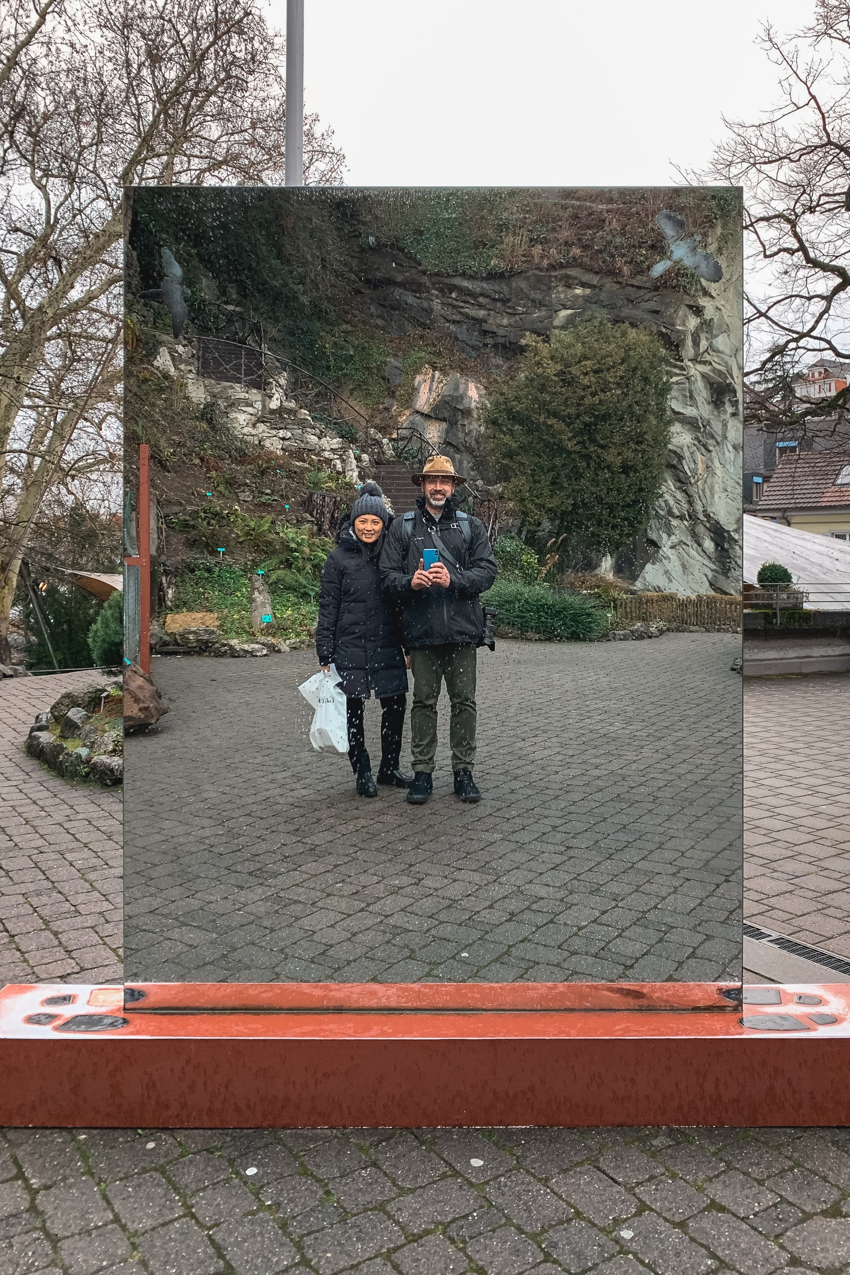 Two people in dark winter coats and hats walk toward the camera on a brick courtyard, framed within a large mounted photograph showing a moss-covered stone structure and bare trees behind them