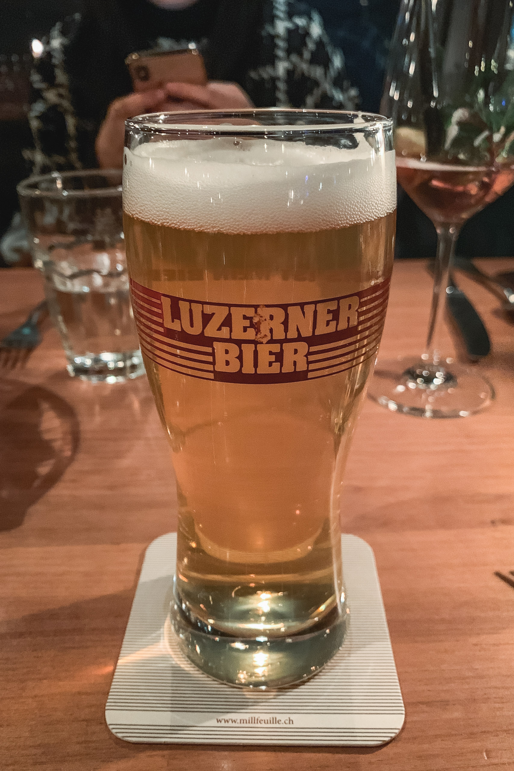 A tall glass of Luzerner Bier with a creamy foam head sits on a coaster at a wooden table, surrounded by cocktail glasses and blurred patrons in warm indoor lighting