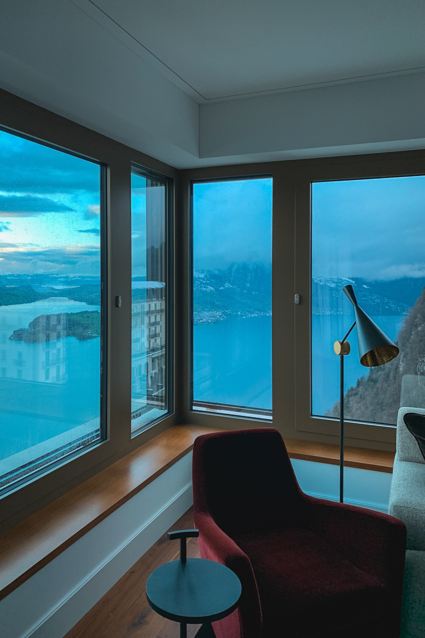 A modern high-rise interior features expansive floor-to-ceiling windows overlooking a blue alpine lake and mountainous landscape at twilight, with a burgundy armchair and floor lamp in the foreground