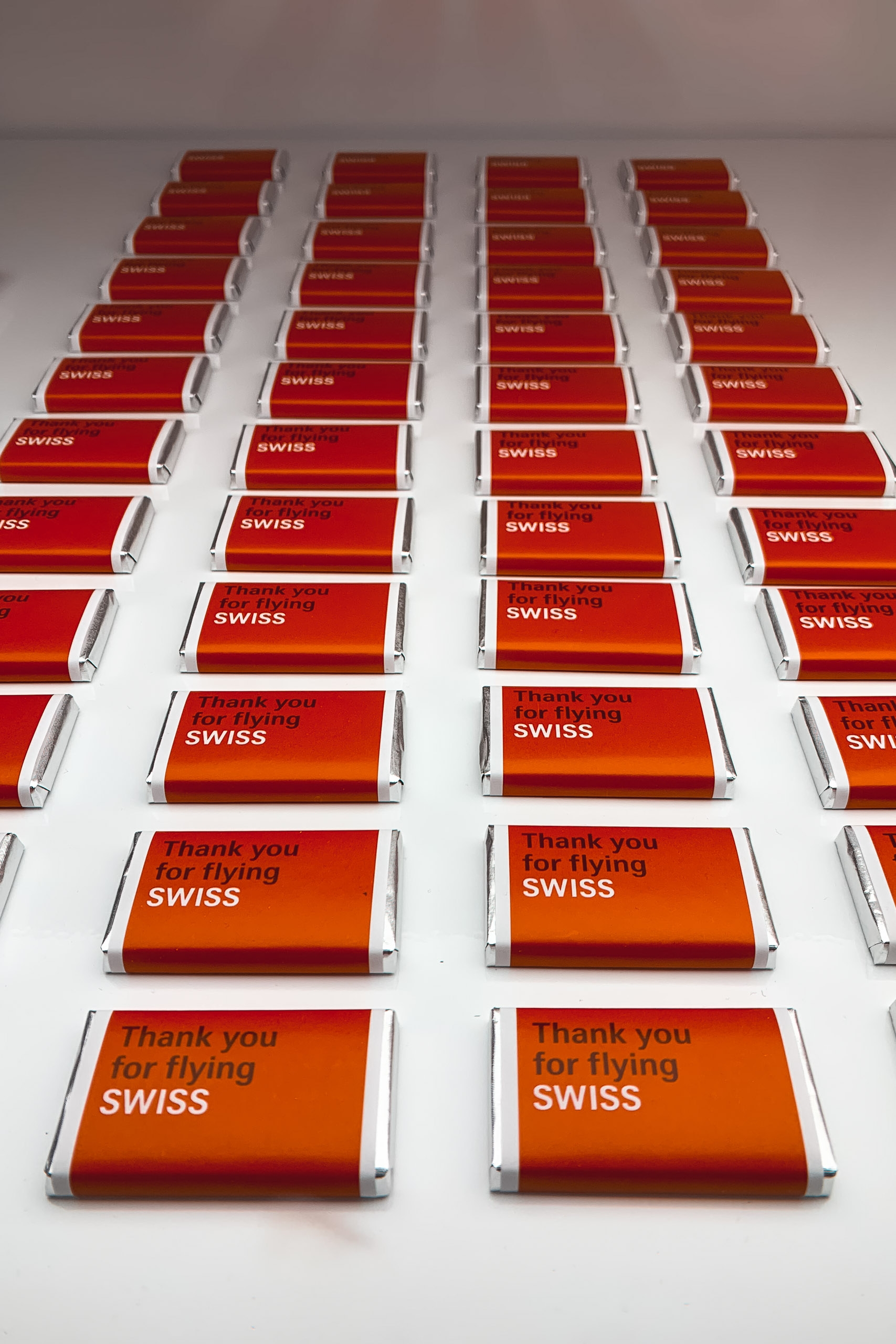 Multiple rows of Swiss airline chocolate bars with red and orange wrappers bearing the "SWISS" logo are arranged in neat columns on a white surface, with "Thank you for flying SWISS" visible on the front packages