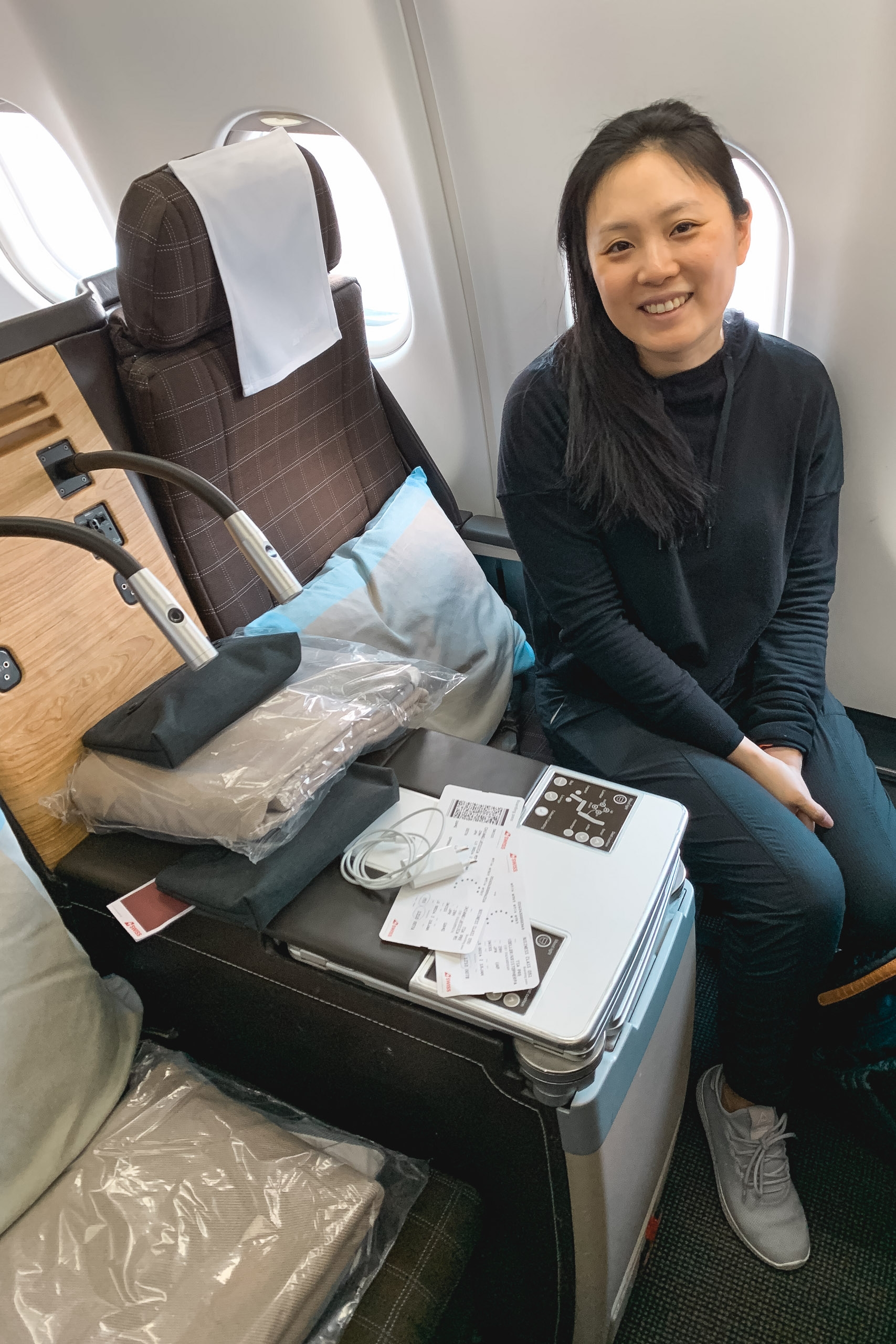 A woman in dark clothing smiles at the camera while seated in an airplane seat surrounded by travel documents, headphones, and packed belongings near a bright cabin window