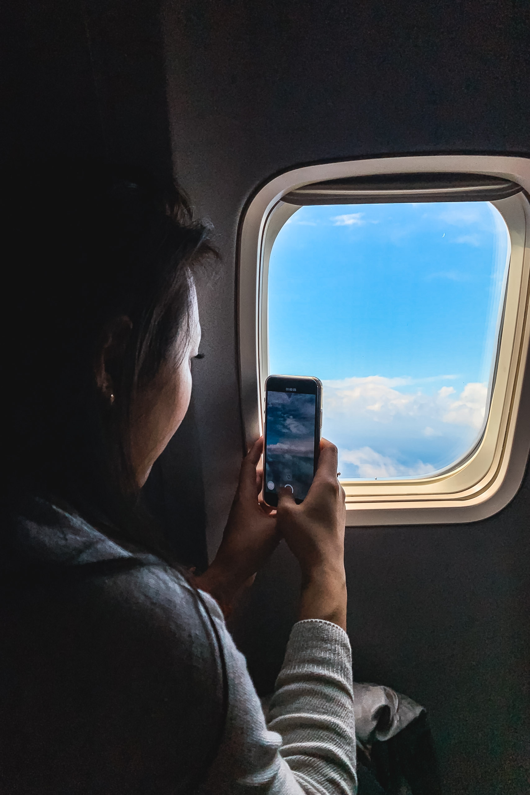A passenger photographs clouds and blue sky through an airplane window, holding a smartphone to capture the aerial view during flight