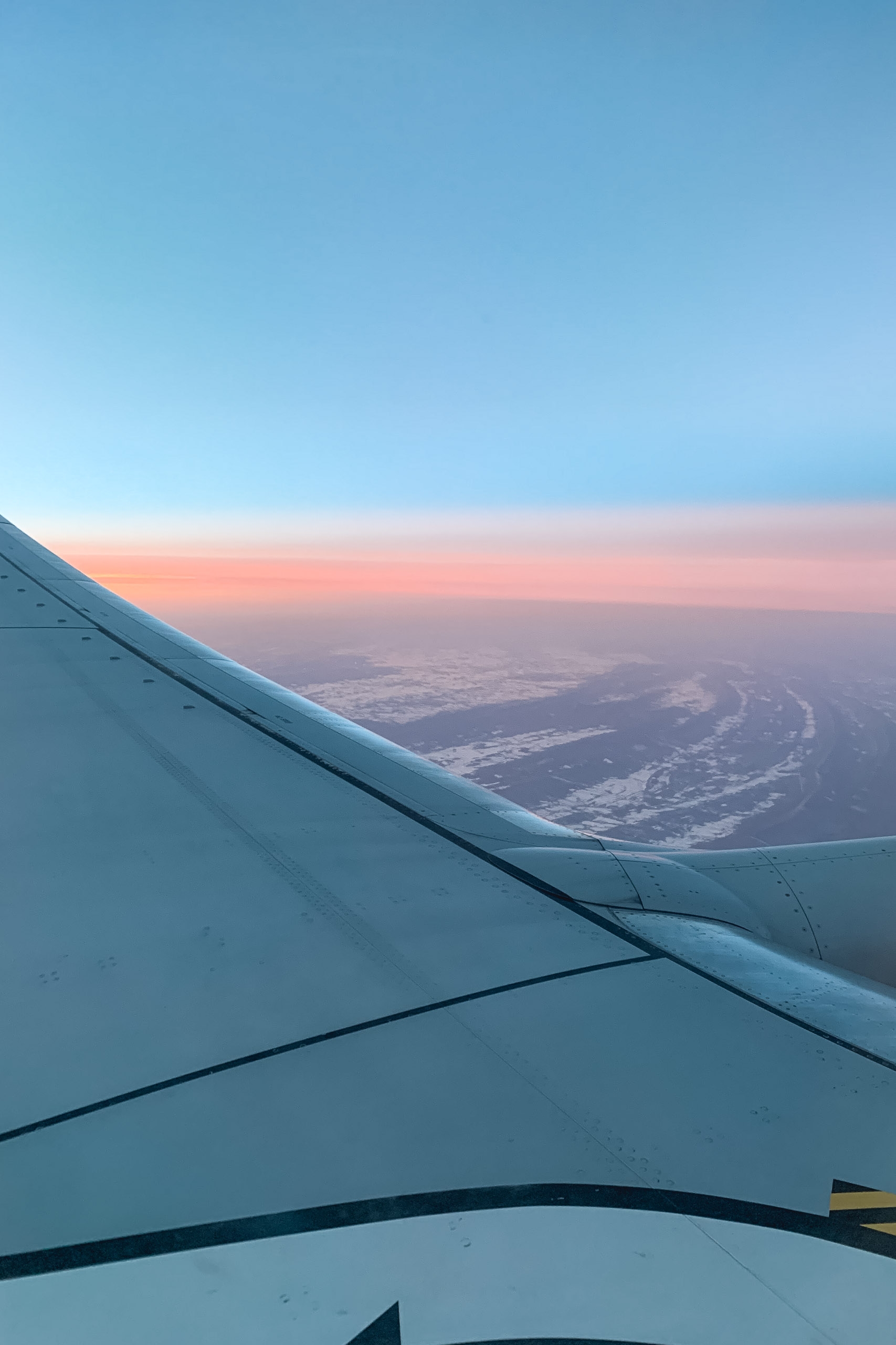 Aircraft wing stretches across the frame during sunrise, revealing a winding river and snowy landscape below beneath a gradient sky transitioning from blue to pink