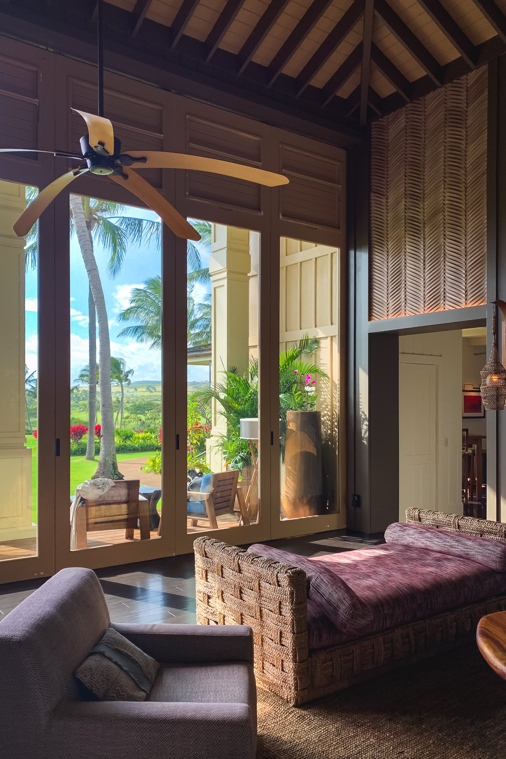 A tropical bedroom with woven furniture and wooden ceiling beams opens through large glass doors to reveal a palm tree-dotted landscape and distant ocean view bathed in warm natural light