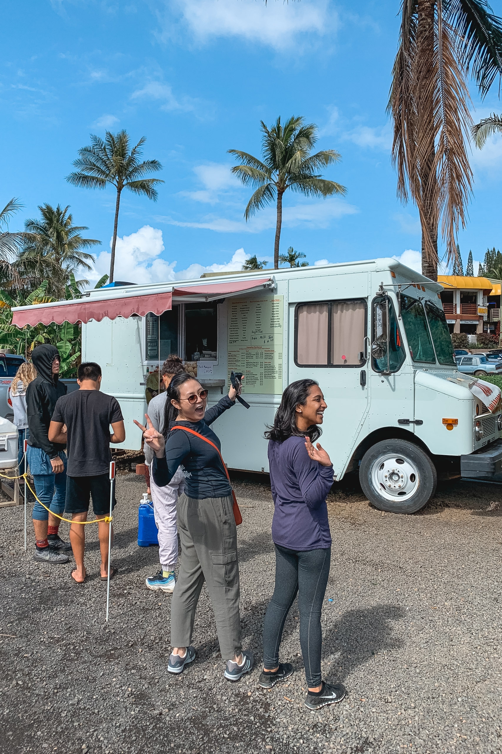 Customers queue at a white food truck with a red awning beneath tall palm trees under bright blue Hawaiian sky