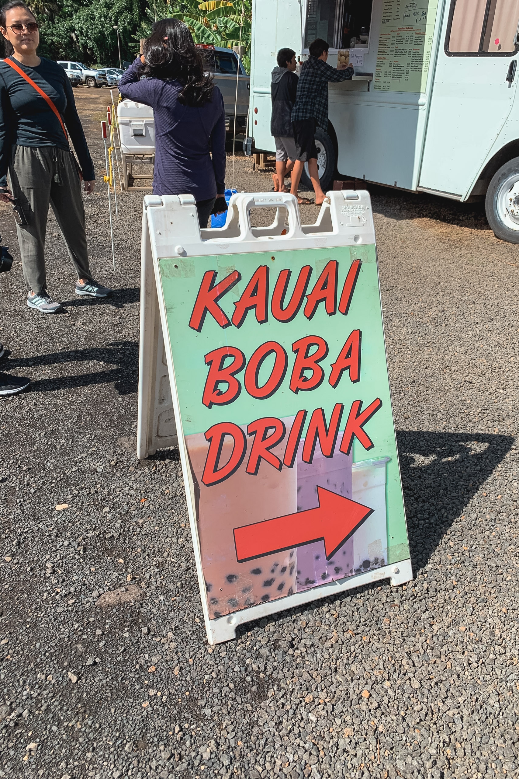 A hand-painted sandwich board advertises Kauai Boba Drink with red lettering and an arrow, positioned on asphalt outside a food truck with customers waiting in line behind it