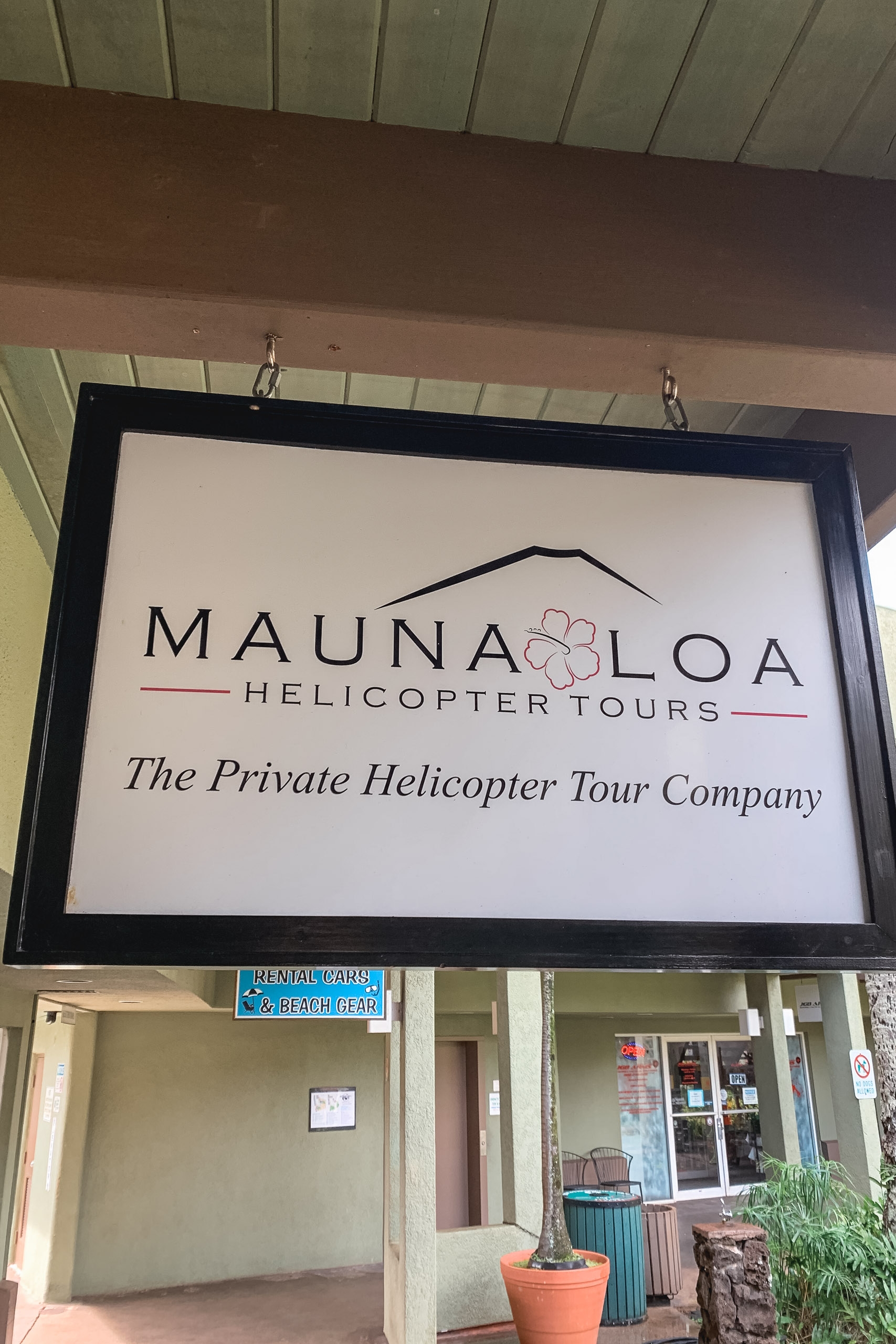 A black-framed business sign for Mauna Loa Helicopter Tours hangs from a wooden overhang, with a potted plant and storefront visible below on an overcast day