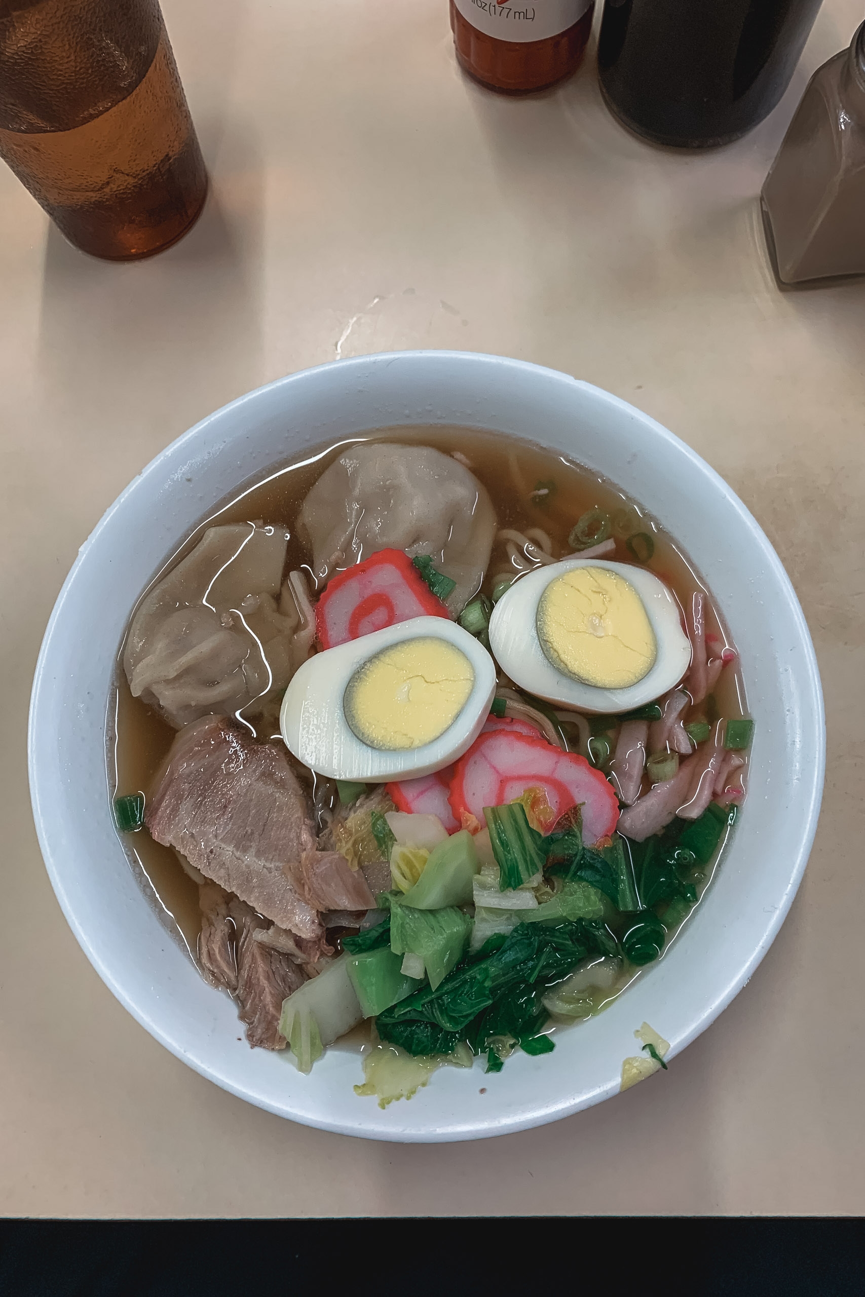 A white bowl of ramen topped with sliced hard-boiled eggs, thin meat slices, pickled ginger, and green vegetables in clear broth, with beer bottles visible in the background on a white table