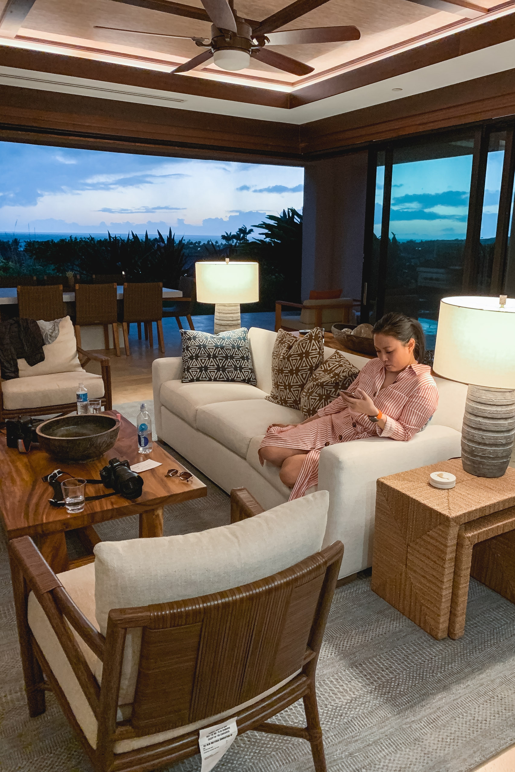 A woman in a pink shirt relaxes on a cream-colored sofa in a modern living room with expansive windows framing a twilight Hawaiian landscape and distant mountains.