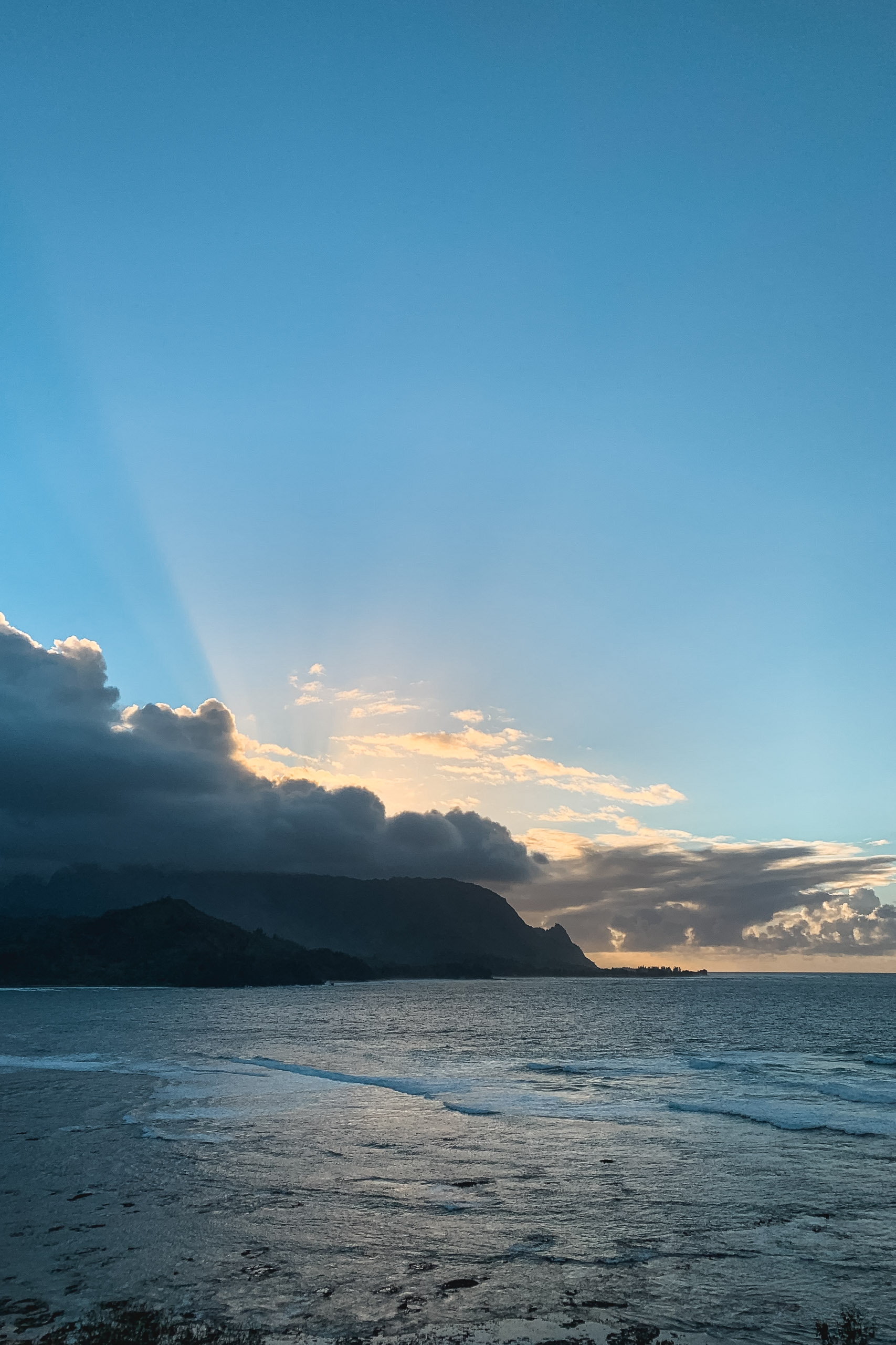 Golden sunlight breaks through dramatic clouds above a silhouetted volcanic coastline, casting rays across calm turquoise waters and wet sand at dusk