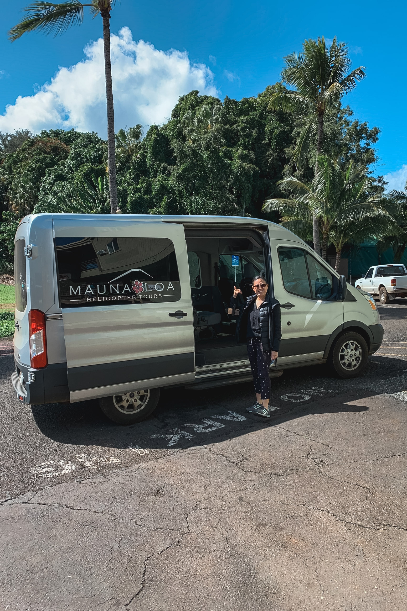 A woman stands beside a white Mauna Loa helicopter tour van in a tropical parking lot under bright blue sky and palm trees