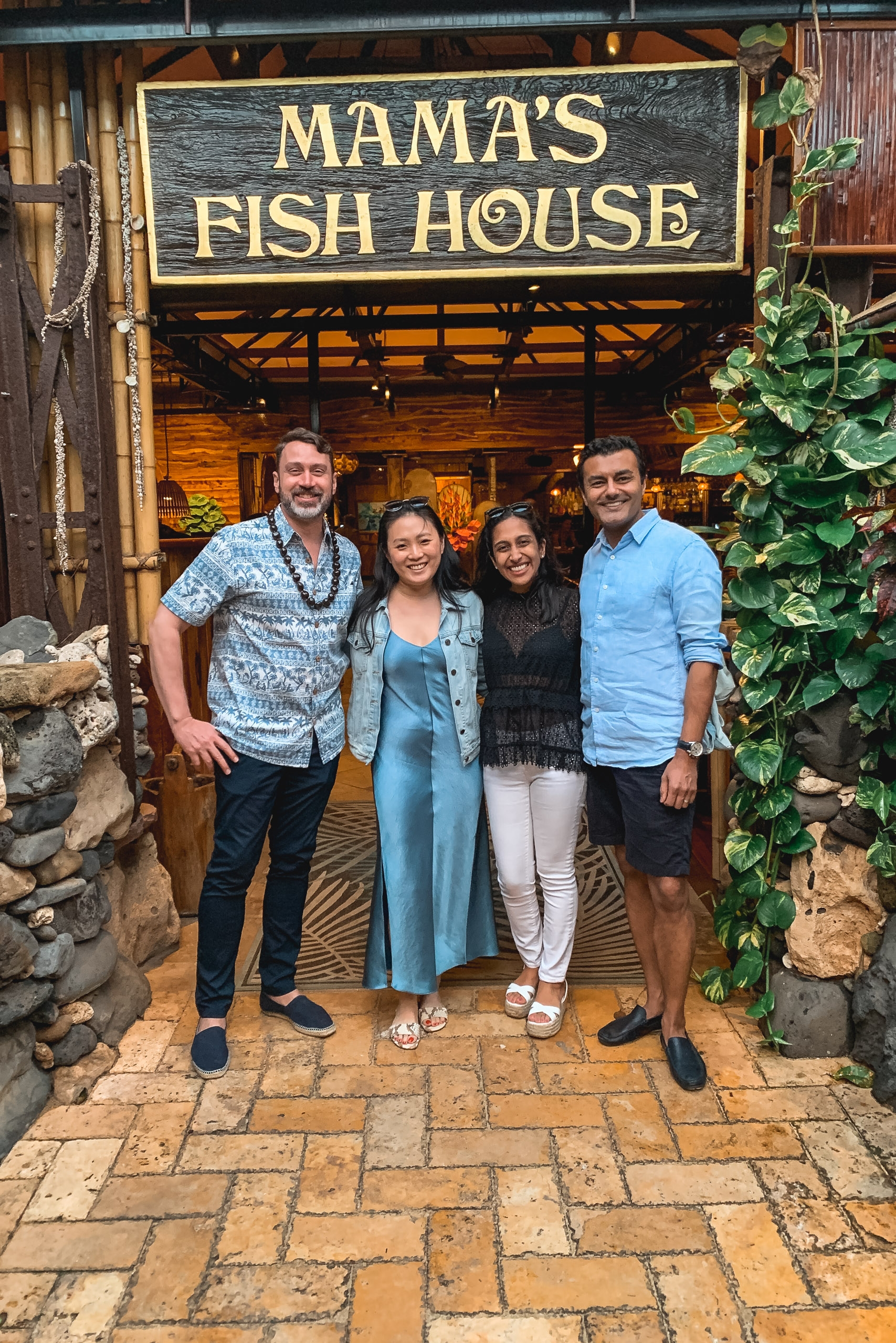 Four people stand smiling beneath the ornate gold-lettered "Mama's Fish House" sign at a rustic wooden restaurant entrance flanked by natural stone and climbing ivy