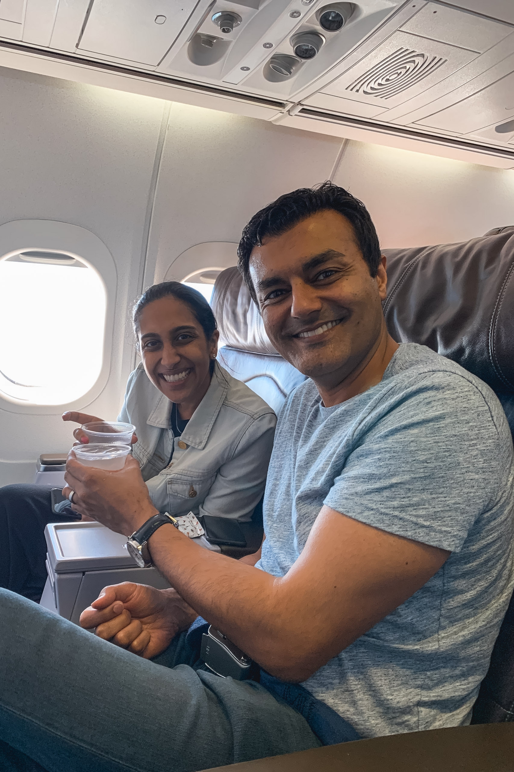 Two smiling passengers sit together in an airplane cabin, holding drinks while bright daylight streams through the window beside them