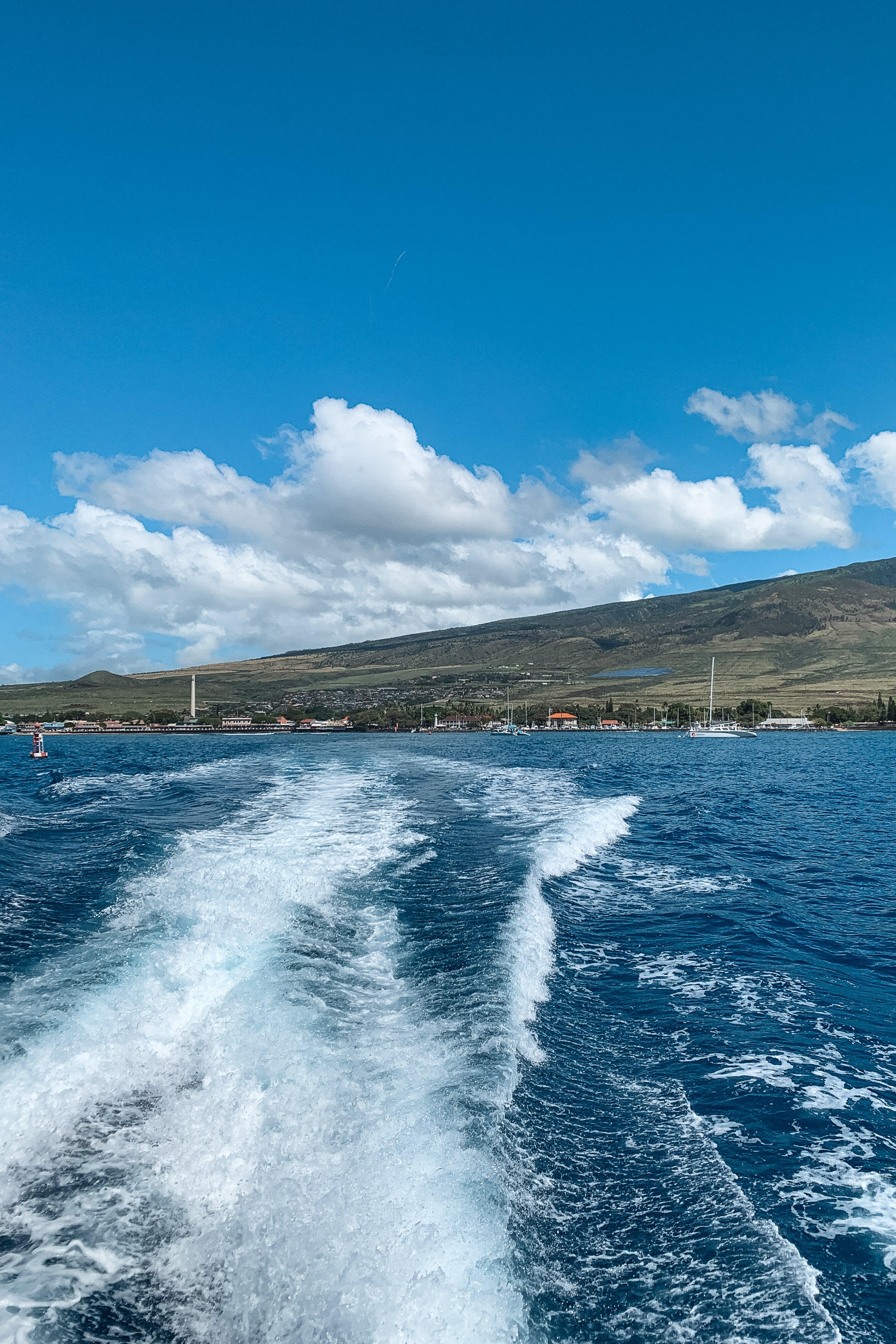 White wake trails stream behind a boat departing a coastal Hawaiian town nestled against a volcanic mountainside under bright blue sky with white cumulus clouds