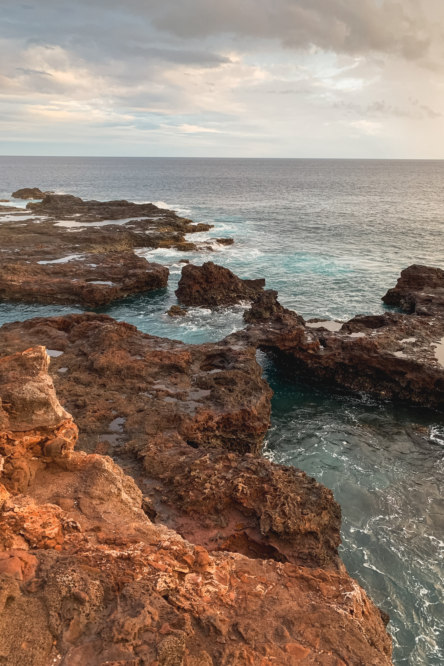 Rocky brown outcrops jut into turquoise ocean waters beneath an overcast sky with soft, diffused light creating a calm coastal seascape
