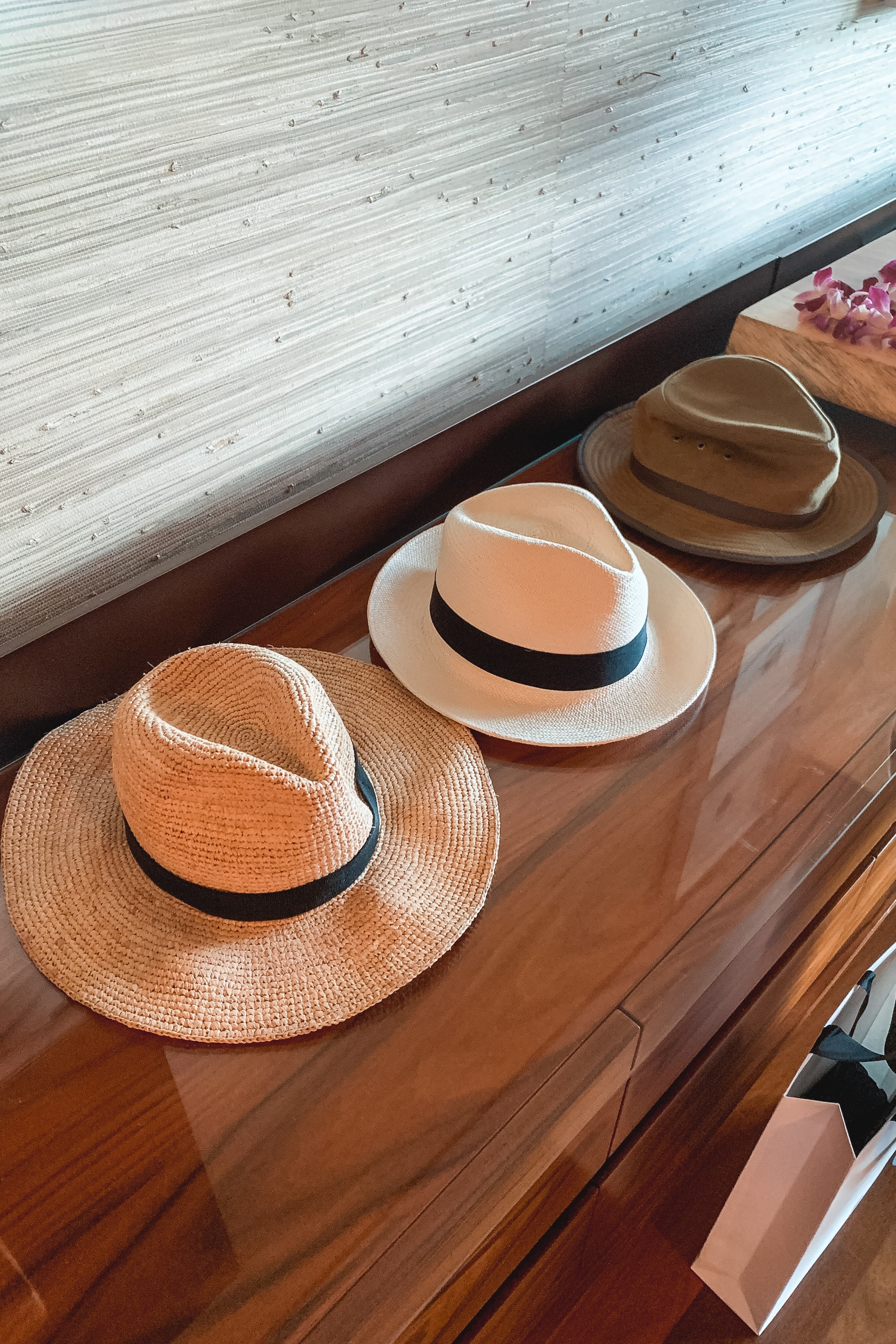 Three straw hats with black bands arranged on a wooden dresser beside a bright window with natural daylight streaming in