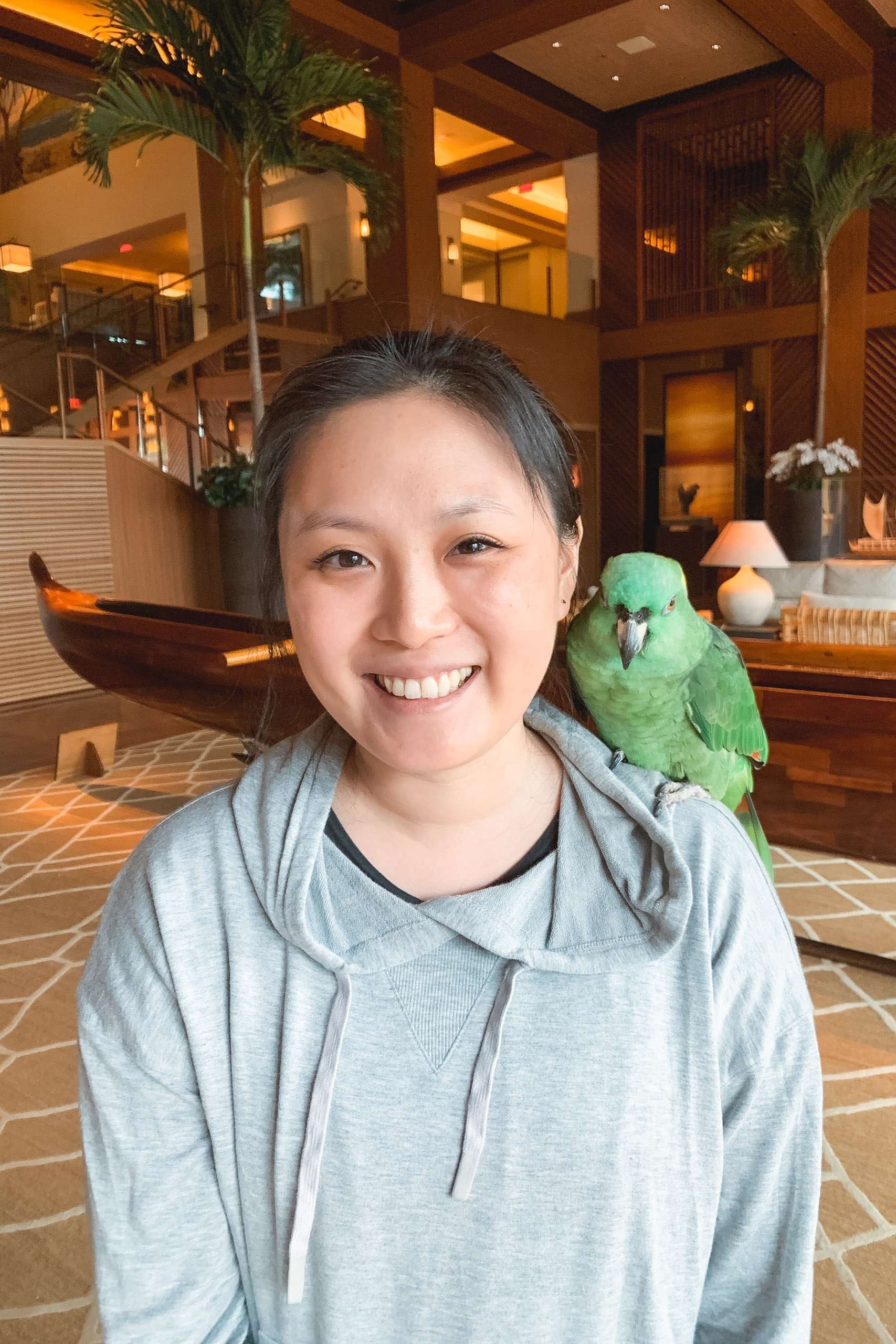 A smiling woman in a gray hoodie poses indoors with a bright green parrot perched on her shoulder, warm wooden architecture and potted plants visible in the luxurious resort lobby behind her.