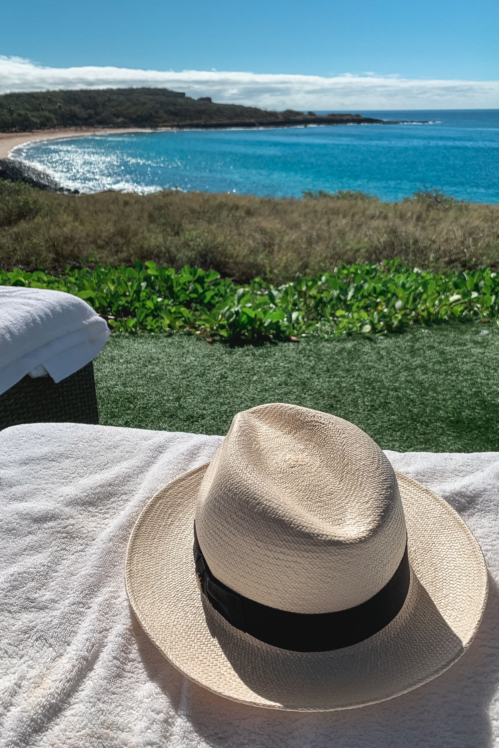 A cream-colored straw hat with a black band rests on white towels overlooking a turquoise bay, with coastal vegetation and forested cliffs visible across the bright blue water under clear skies