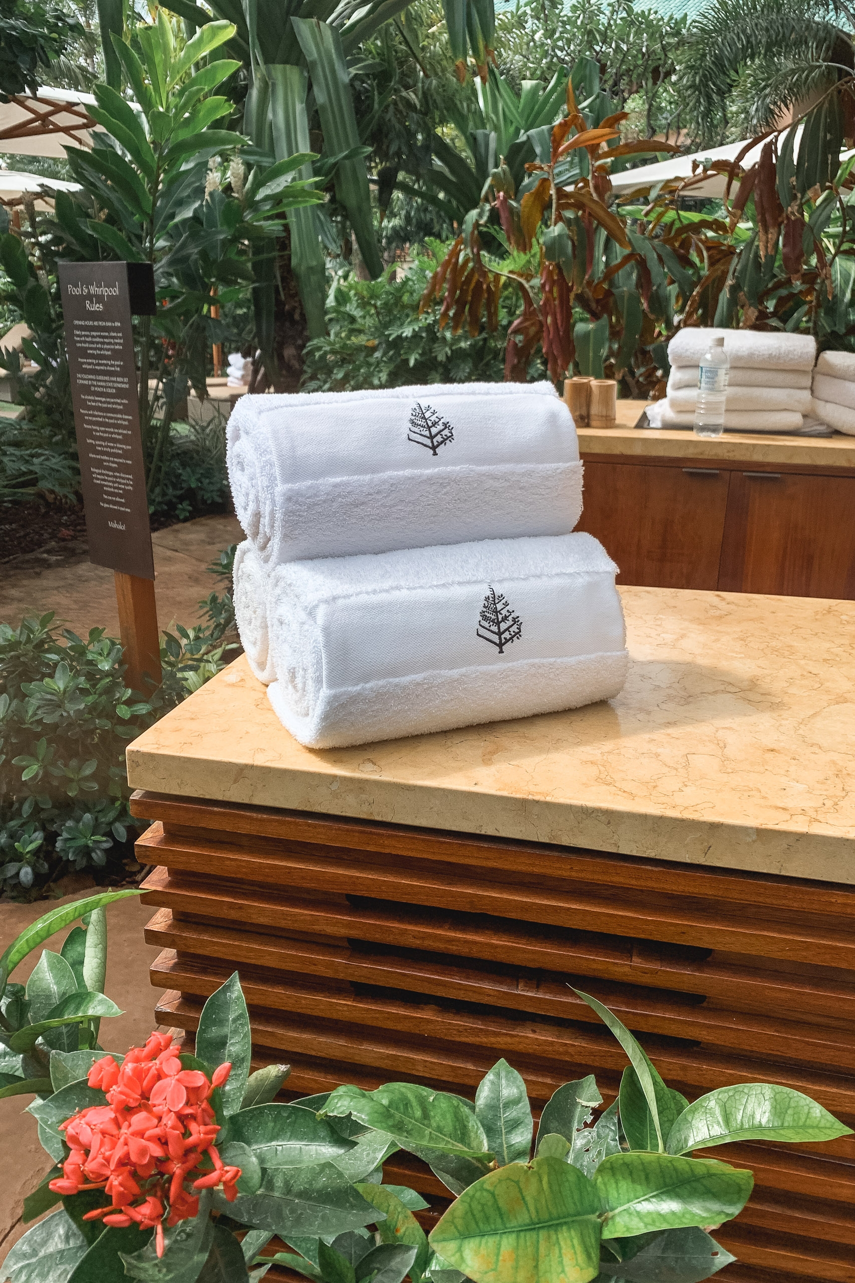 Stacked white rolled towels with embroidered leaf designs sit on a wooden display counter surrounded by lush tropical plants and red flowers in a greenhouse setting