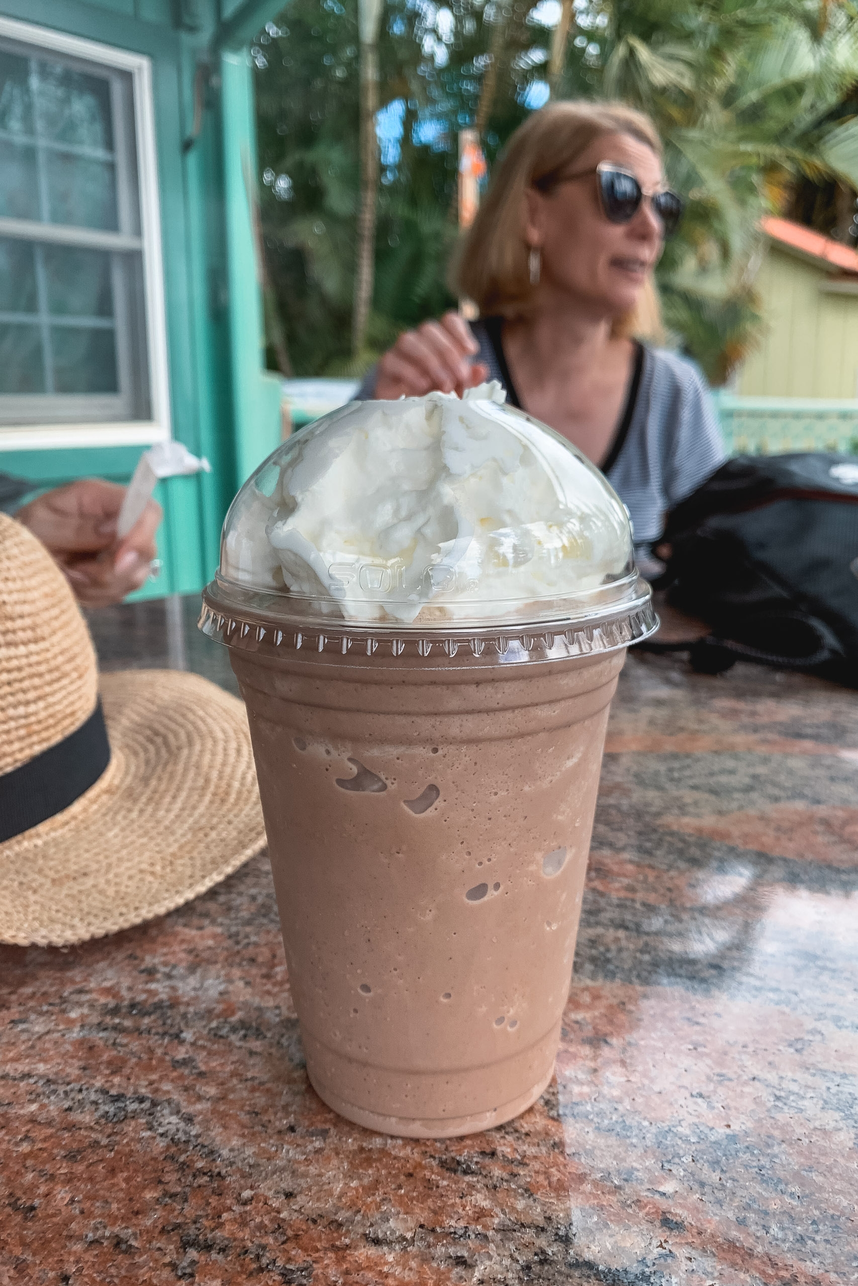 A cold chocolate beverage topped with whipped cream sits on a brick surface while a woman wearing sunglasses relaxes in the background near a turquoise building