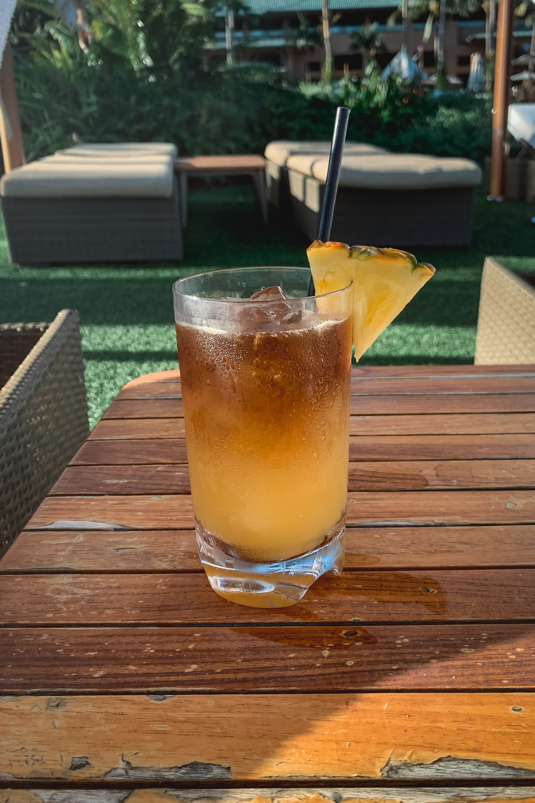 An amber-colored tropical cocktail with ice and a pineapple wedge garnish sits on a wooden table in a sunlit garden patio setting with lounge chairs and lush greenery