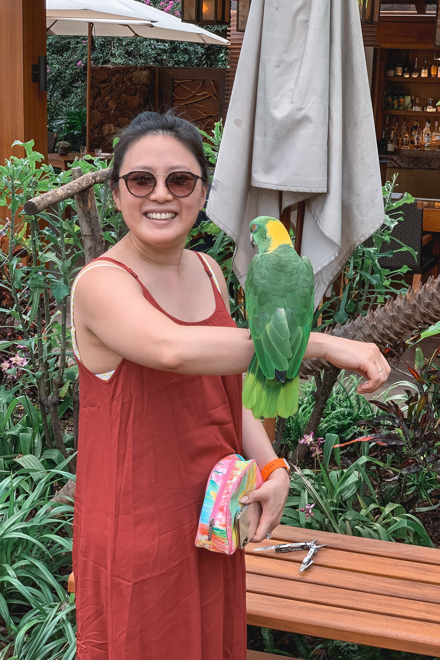 Woman in red dress and glasses smiles while holding a green and yellow parrot on her arm in a lush tropical garden setting