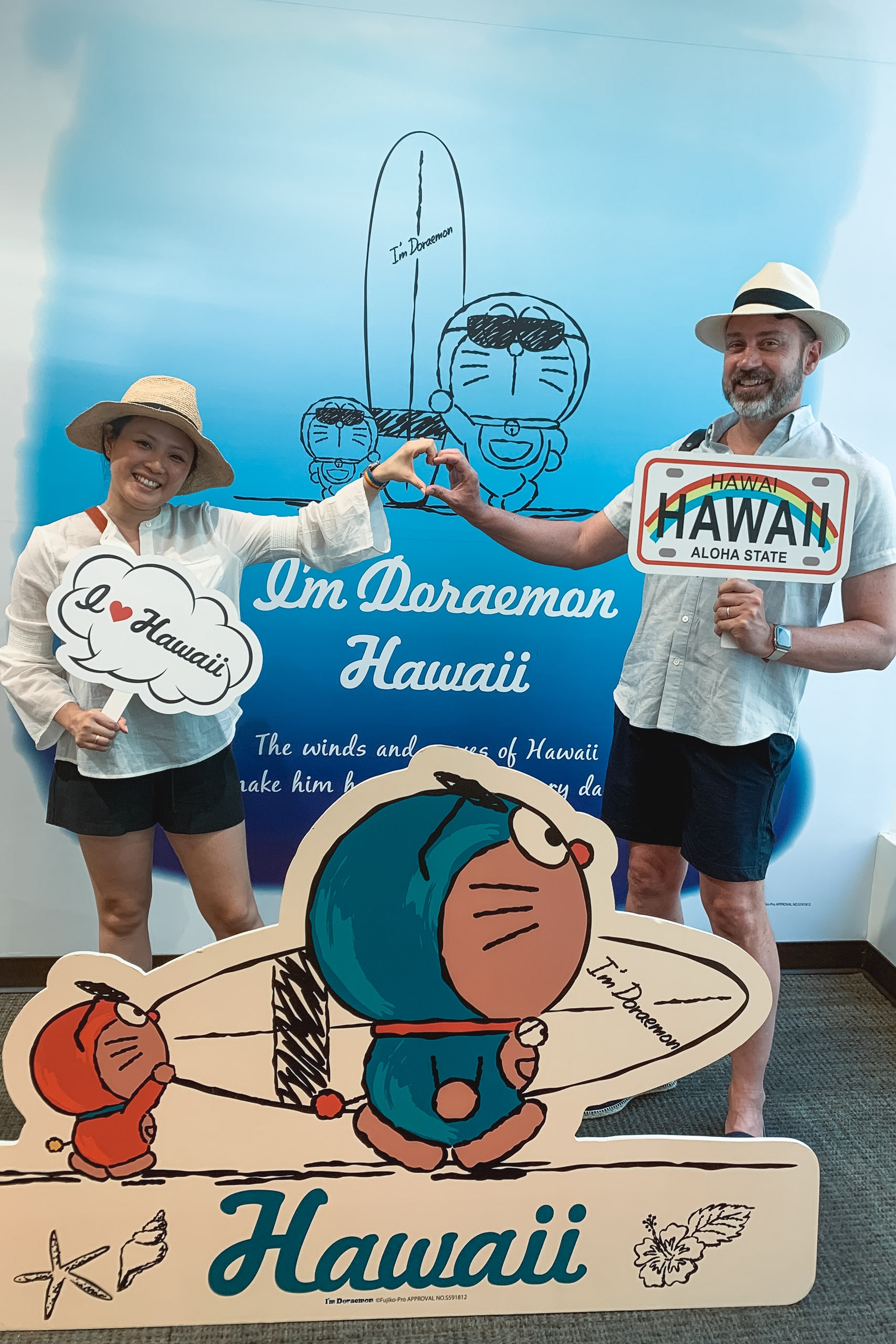 Two tourists in straw hats pose with oversized cardboard cutouts of Doraemon characters against a blue Hawaiian-themed backdrop, making hand gestures and holding props