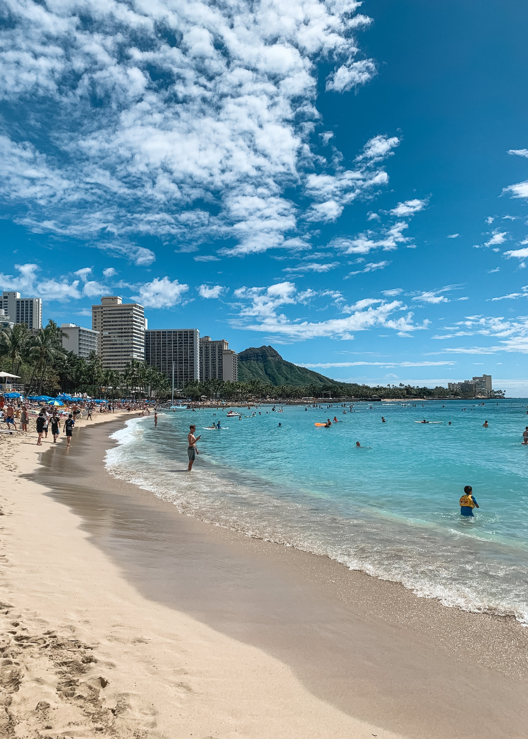 Beachgoers swim in turquoise waters along a sandy shore with high-rise hotels, palm trees, and a verdant volcanic mountain backdrop under bright blue sky