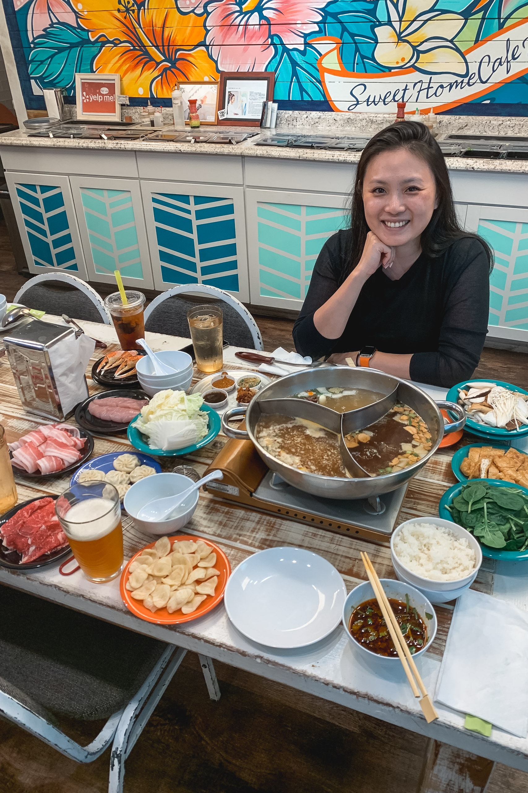 A woman smiles at a hot pot dining table laden with raw meats, vegetables, and broths, with colorful floral mural and turquoise geometric patterns visible behind her