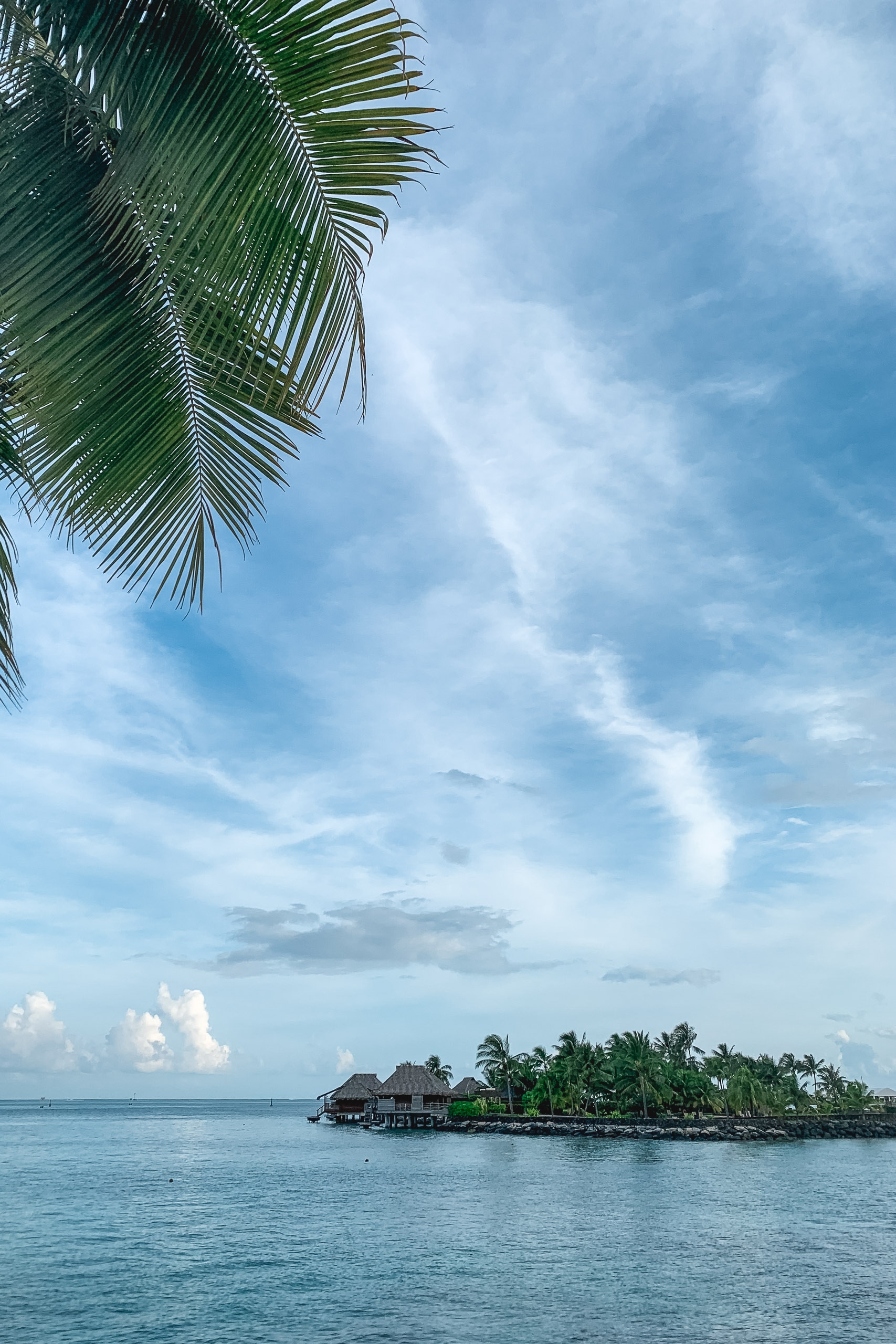 Palm fronds frame a turquoise lagoon view with a small island dotted with thatched bungalows beneath a bright, cloud-streaked sky