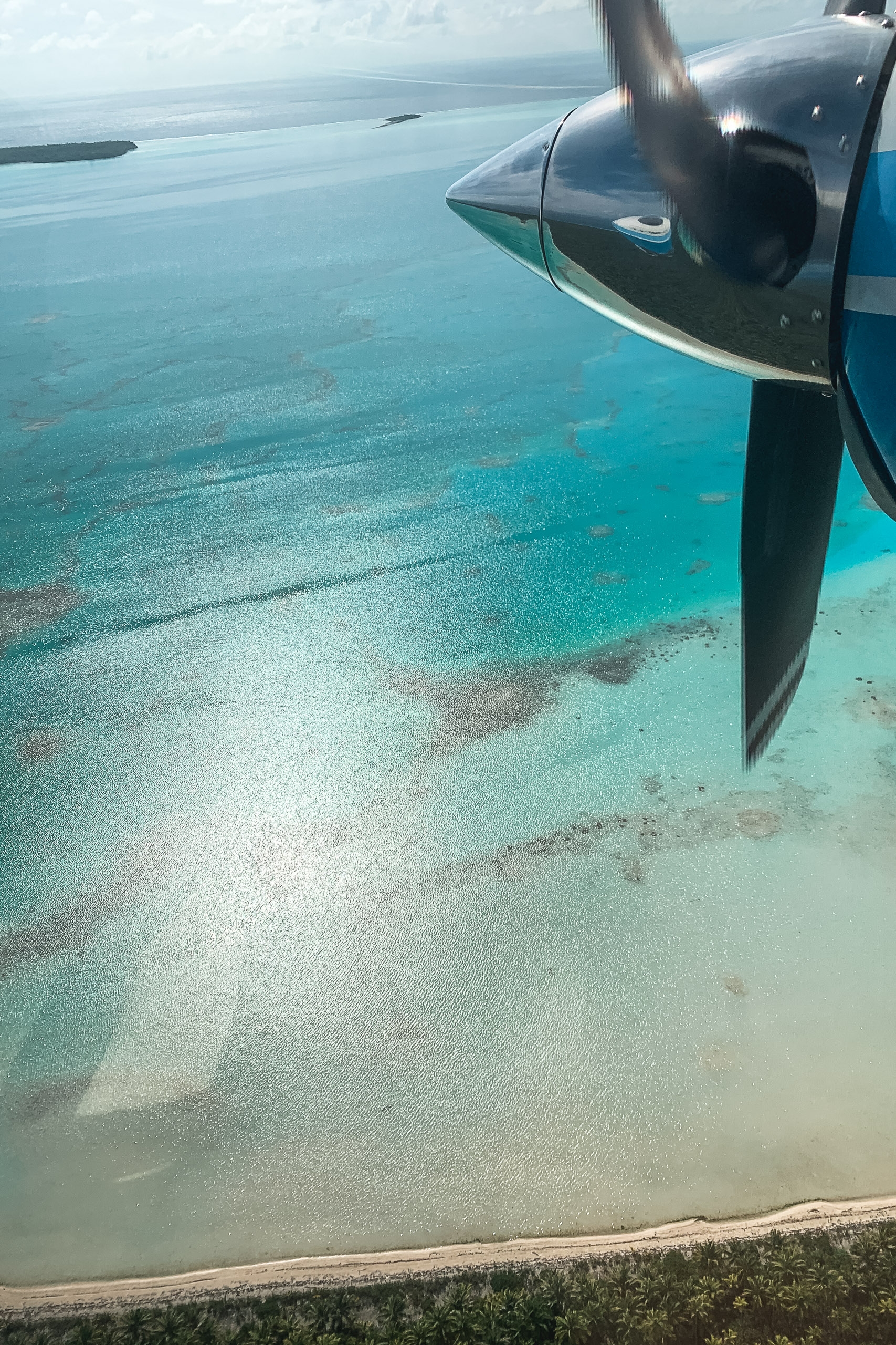 A propeller and engine nacelle of a small aircraft frame turquoise tropical waters and sandy shallows below during a scenic aerial flight over a Pacific island atoll.
