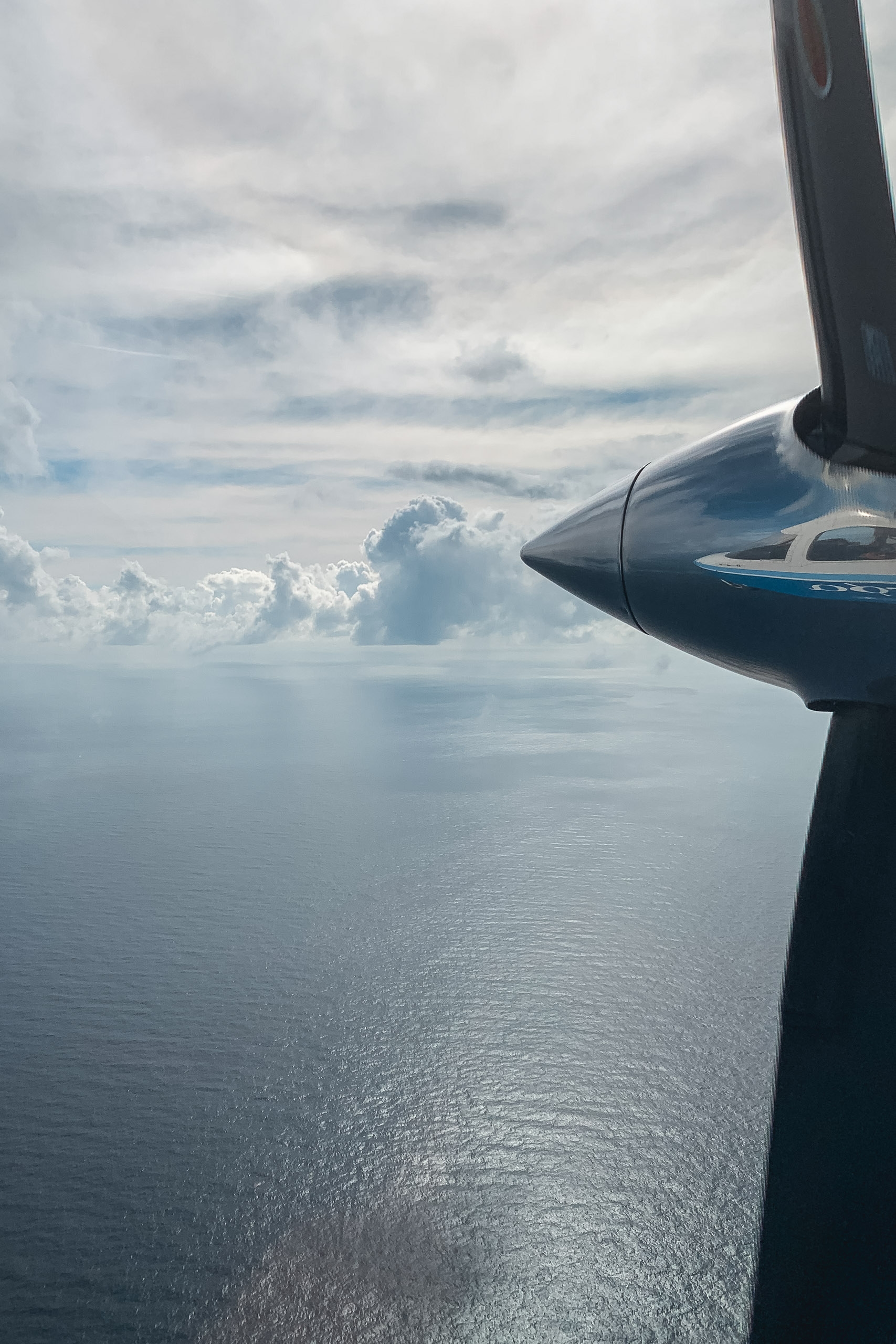Aircraft propeller and engine visible in foreground as small plane flies above calm ocean waters with towering white cumulus clouds scattered across overcast sky