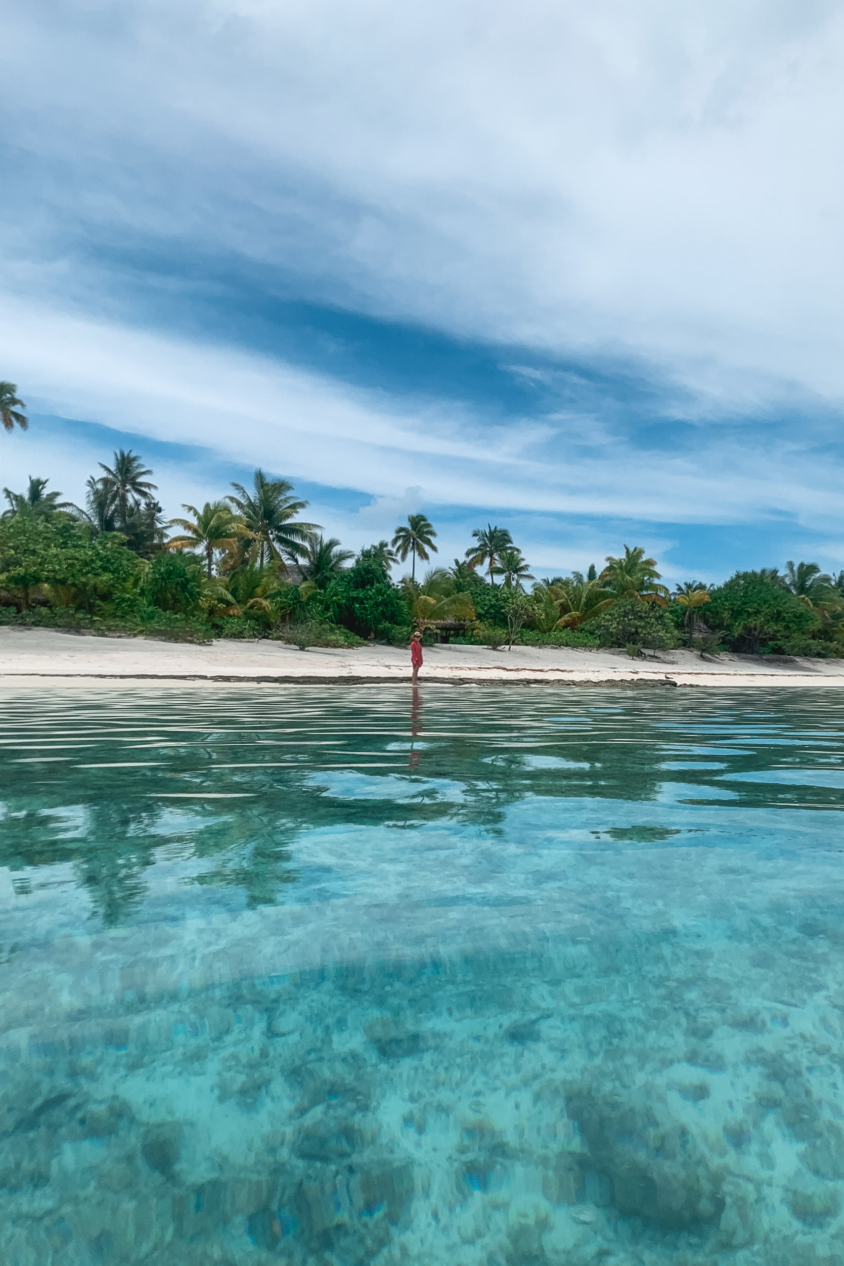 A solitary figure stands in shallow turquoise water off a white sand beach lined with palm trees under a partly cloudy sky