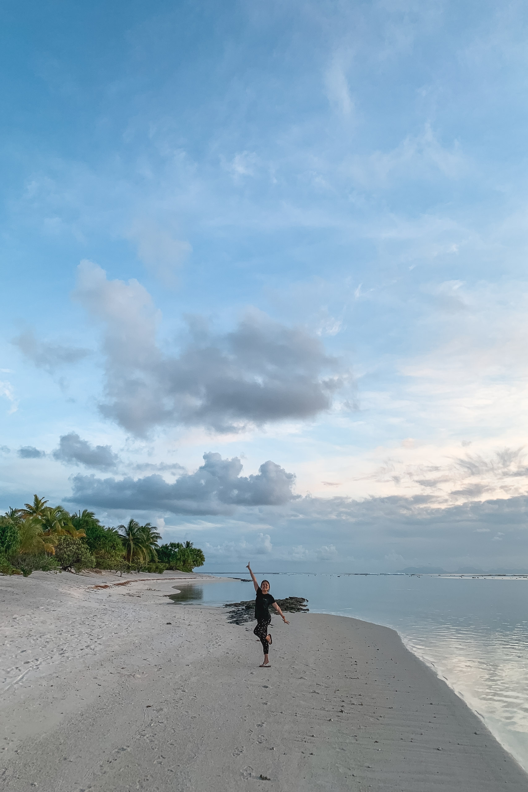 A surfer in a wetsuit walks along a pristine white sand beach toward calm turquoise waters beneath a partly cloudy sky, with palm trees visible along the shoreline