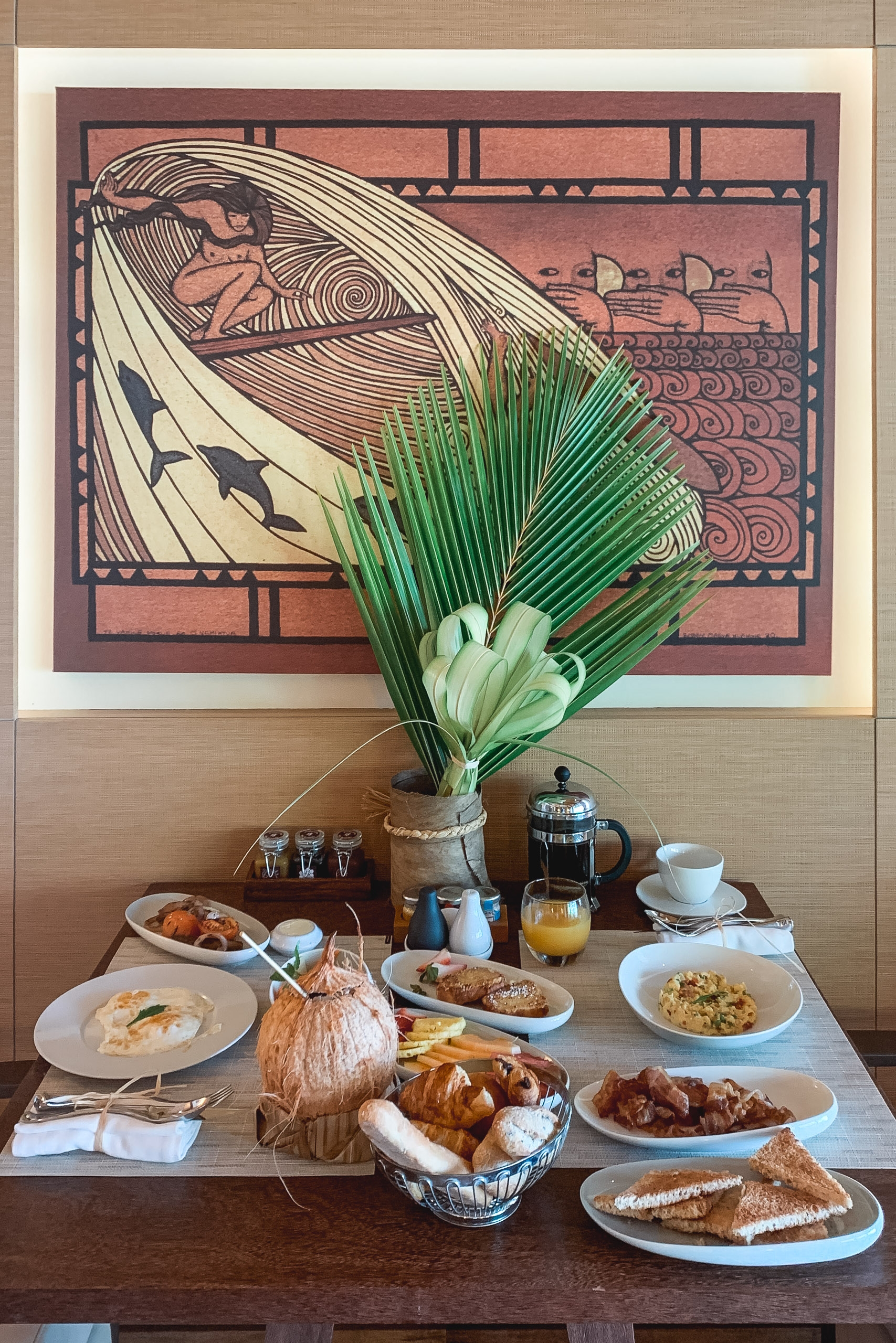 A breakfast spread with pastries, fresh fruit, and dishes arranged on a table beneath a stylized Polynesian artwork and tall green palm fronds in a vase