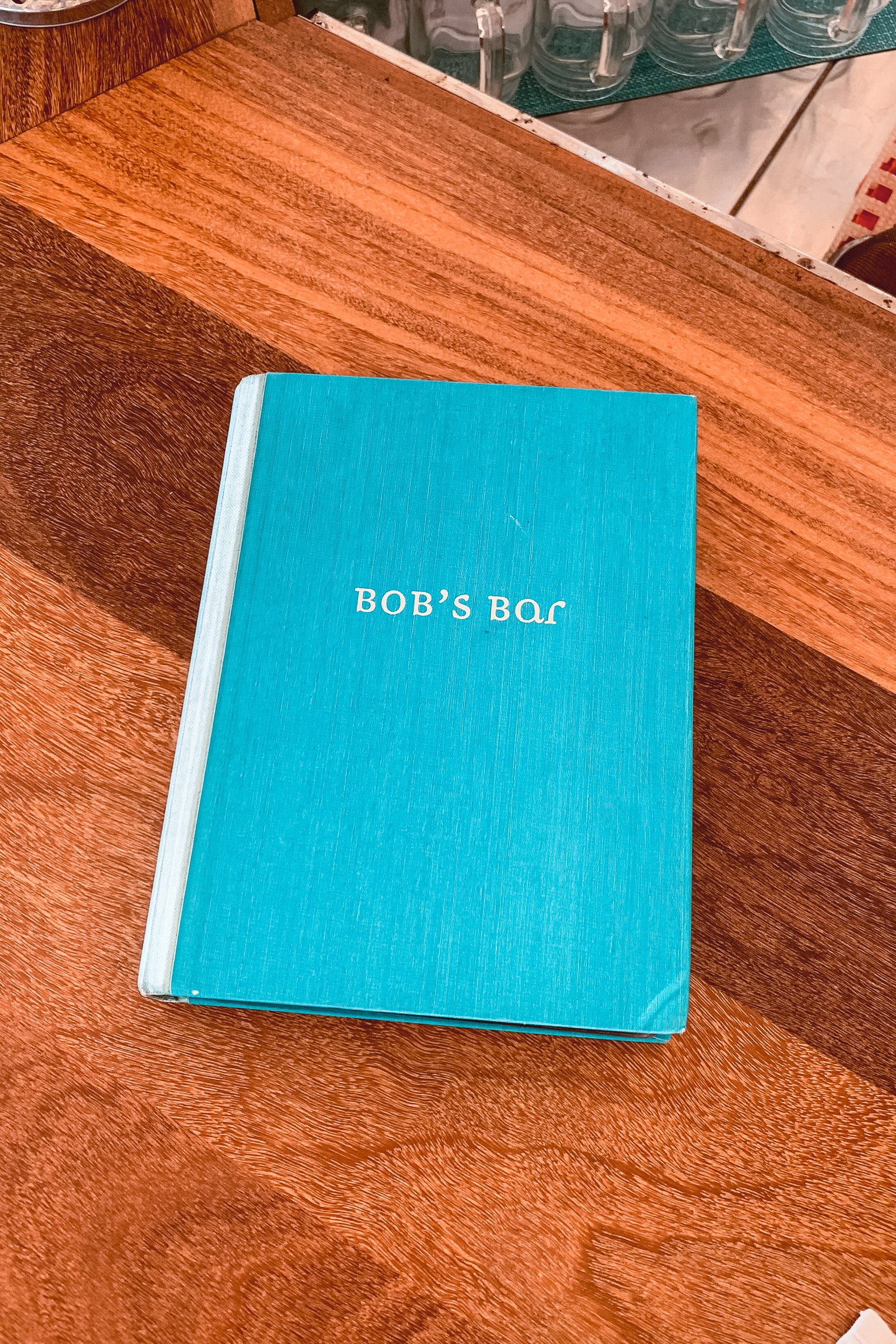 A bright turquoise menu labeled "Bob's Bar" rests on warm wooden grain surface with glassware visible in the soft background
