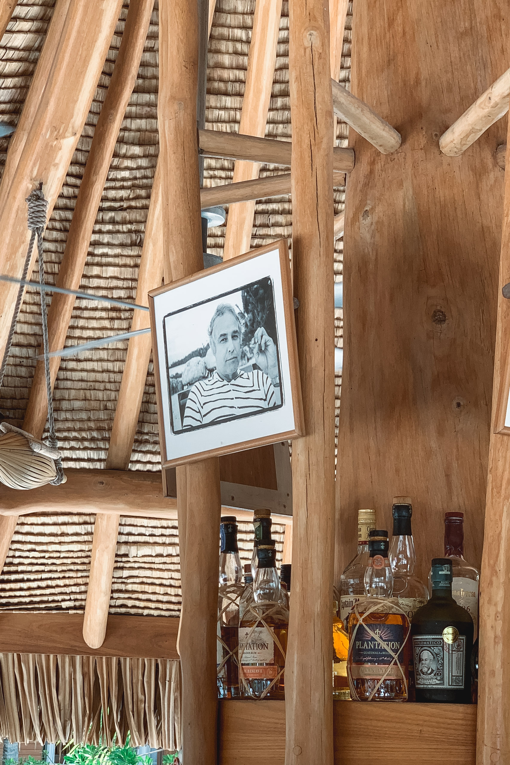 Wooden interior of a tropical establishment displays various spirits bottles on a shelf beneath a black-and-white portrait photograph, with woven palm fronds and wooden beams overhead