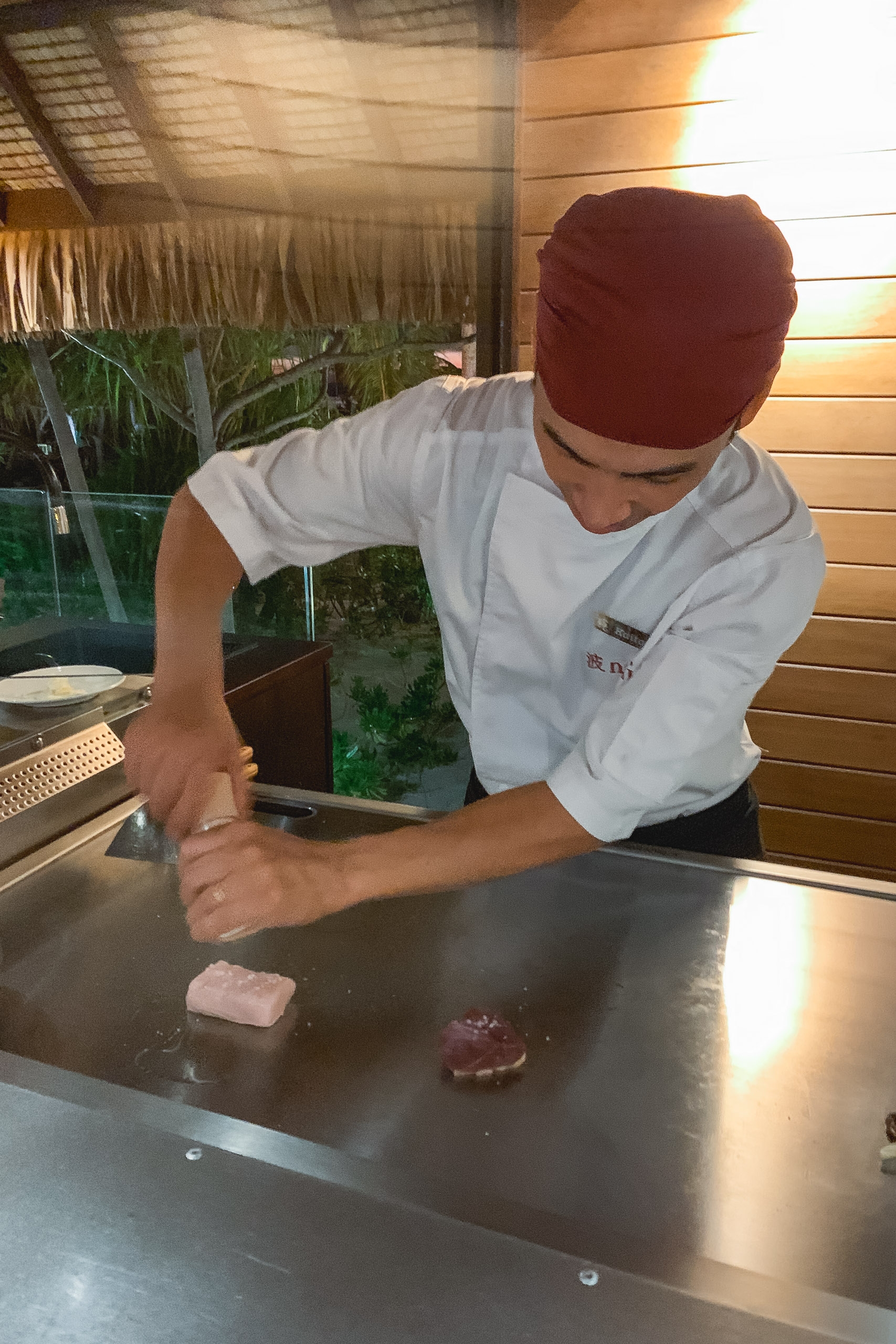 A chef wearing a burgundy headband and white chef's coat prepares food on a stainless steel counter in bright, warm sunlight filtering through wooden slats