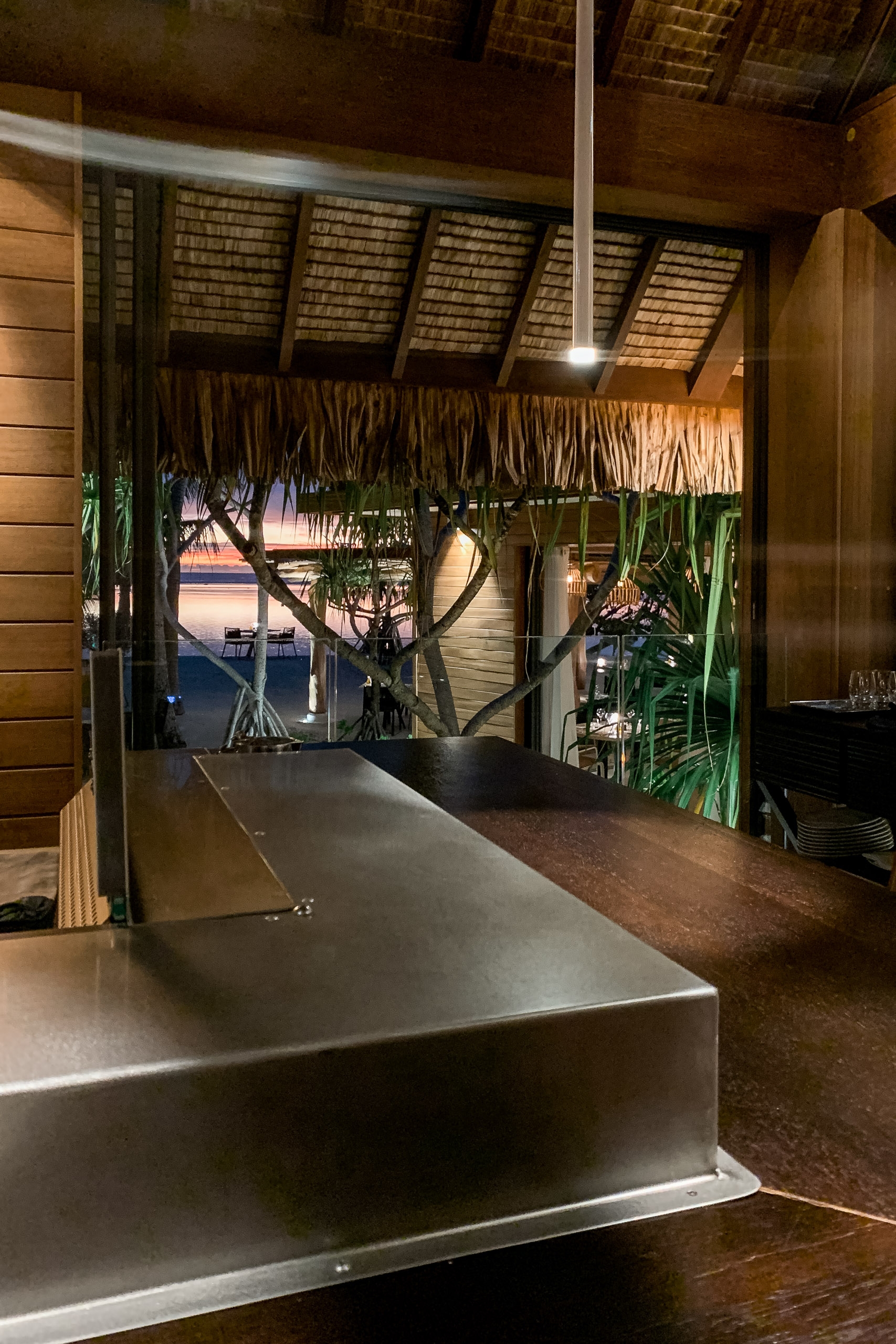 A modern metallic counter extends through a wooden interior space, framing a twilight view of tropical palms and water visible through large glass doors with woven screens overhead
