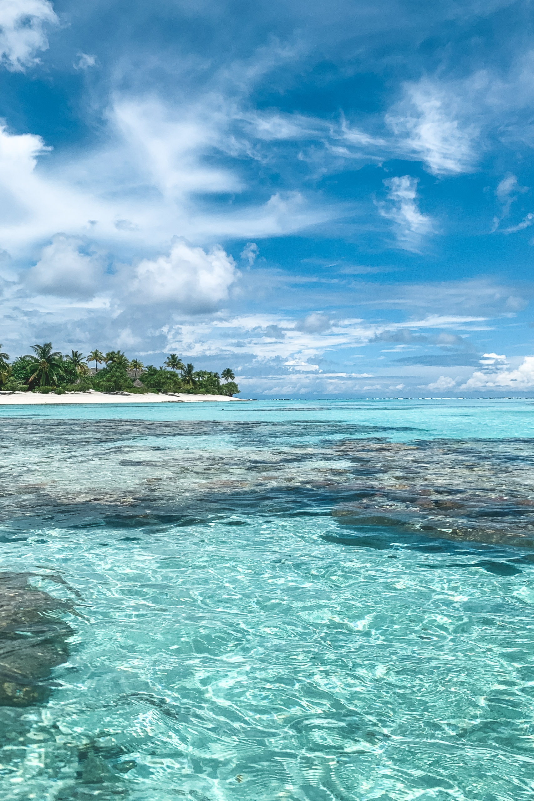 Shallow turquoise lagoon waters with rippling patterns stretch toward a distant palm-fringed island under a bright blue sky scattered with white clouds