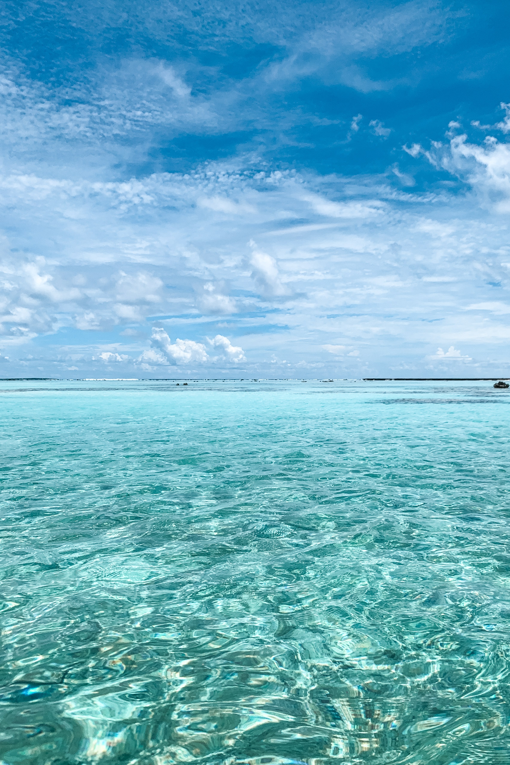 Crystal-clear turquoise lagoon water stretches toward a distant horizon beneath a bright blue sky dotted with white clouds and small boats