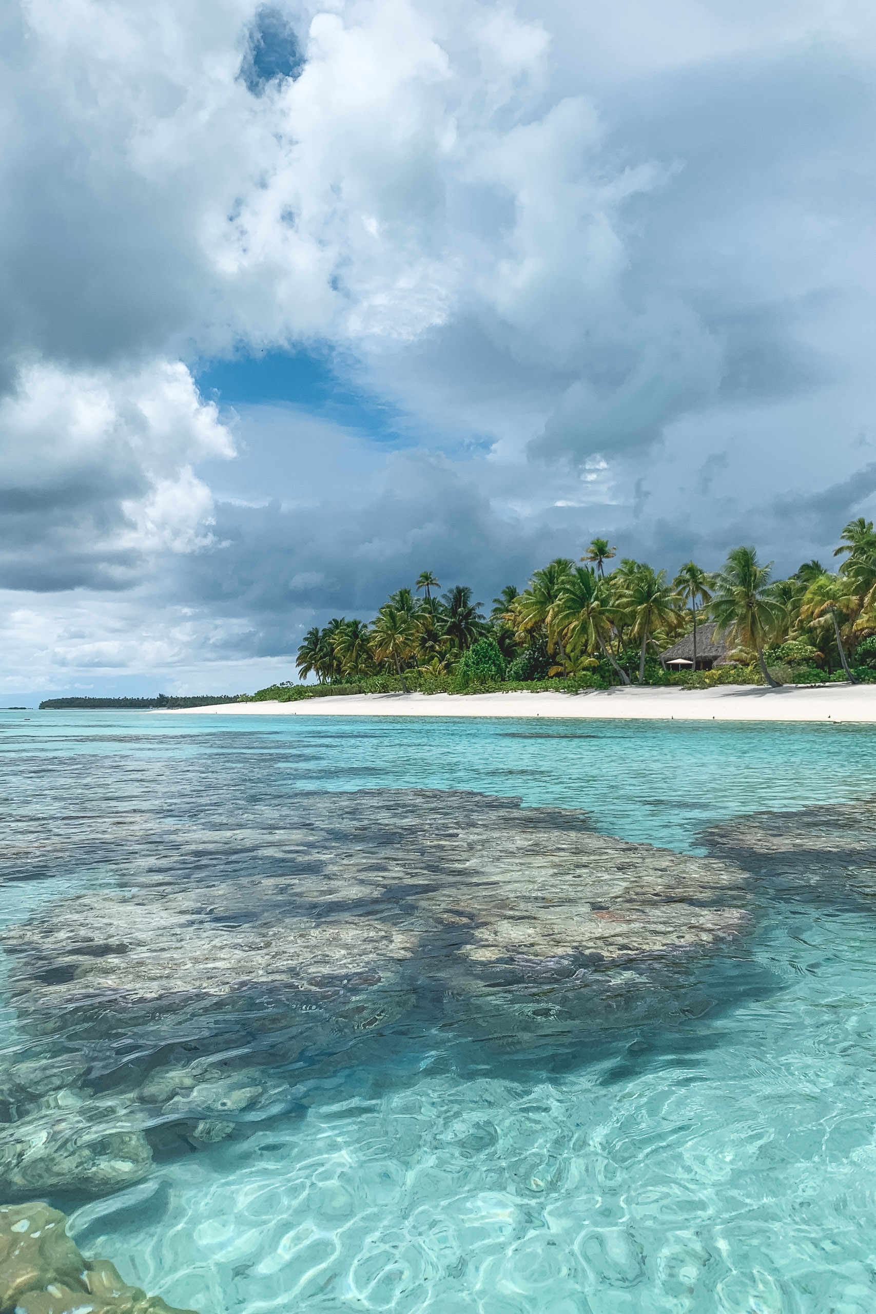 Turquoise shallow waters reveal sandy bottom and coral formations before a pristine white-sand beach lined with coconut palms beneath dramatic cumulus clouds