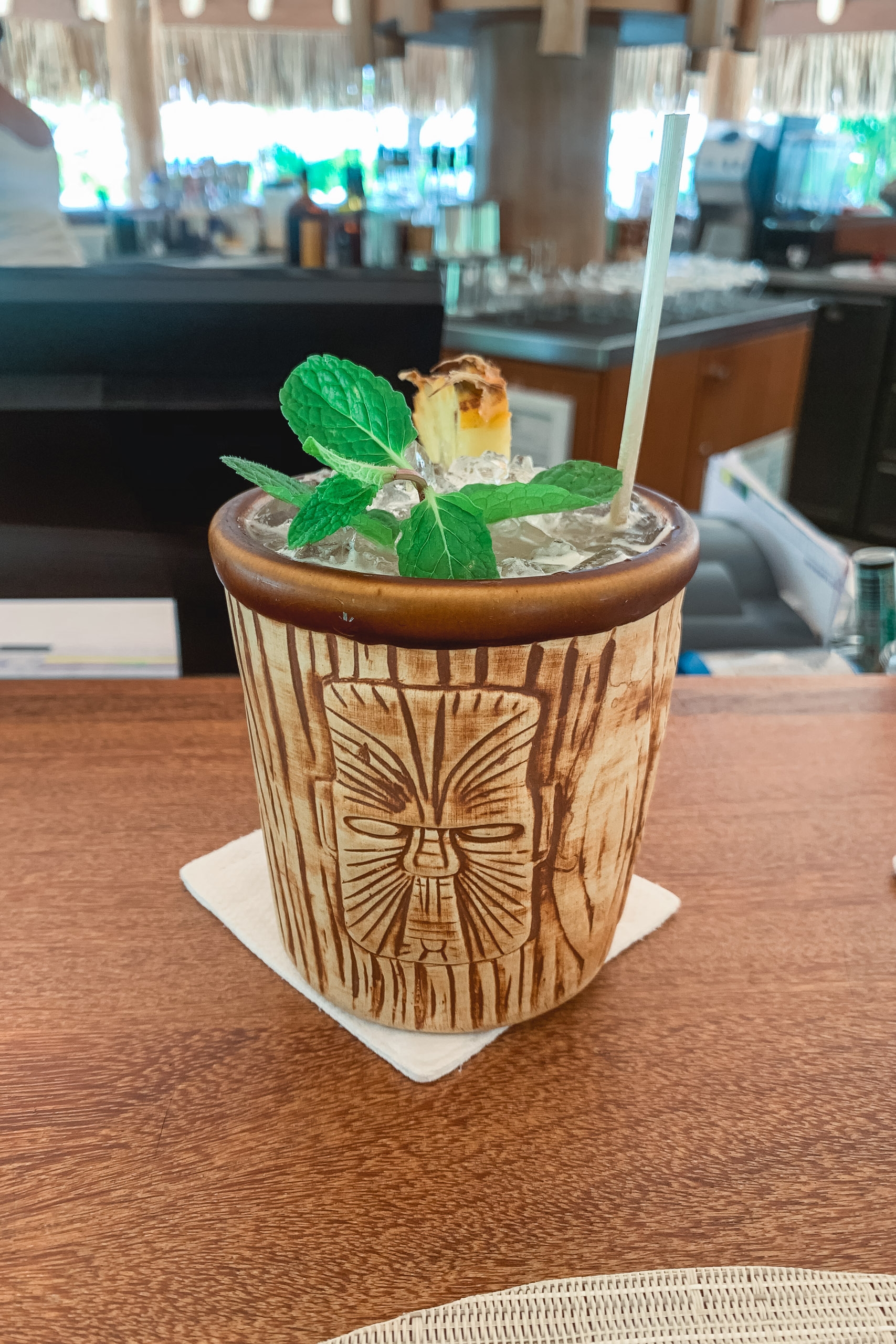 A tropical cocktail in a carved wooden tiki cup sits on a bar counter, garnished with fresh mint and a dried fruit slice, with an out-of-focus resort bar interior behind it.