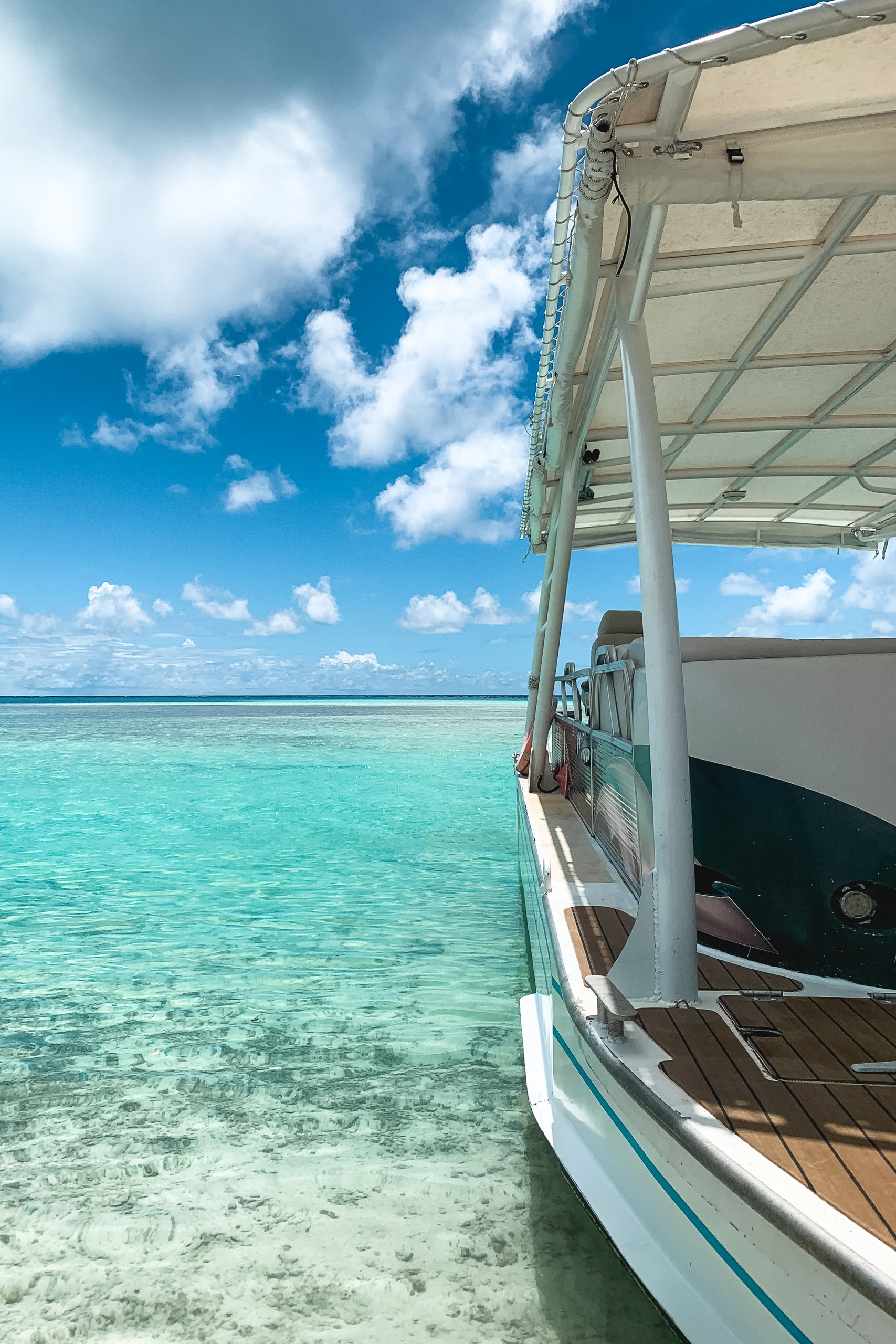 A white boat with canvas canopy floats in crystalline turquoise water under a bright blue sky with white clouds, revealing sandy bottom beneath the shallow tropical lagoon.