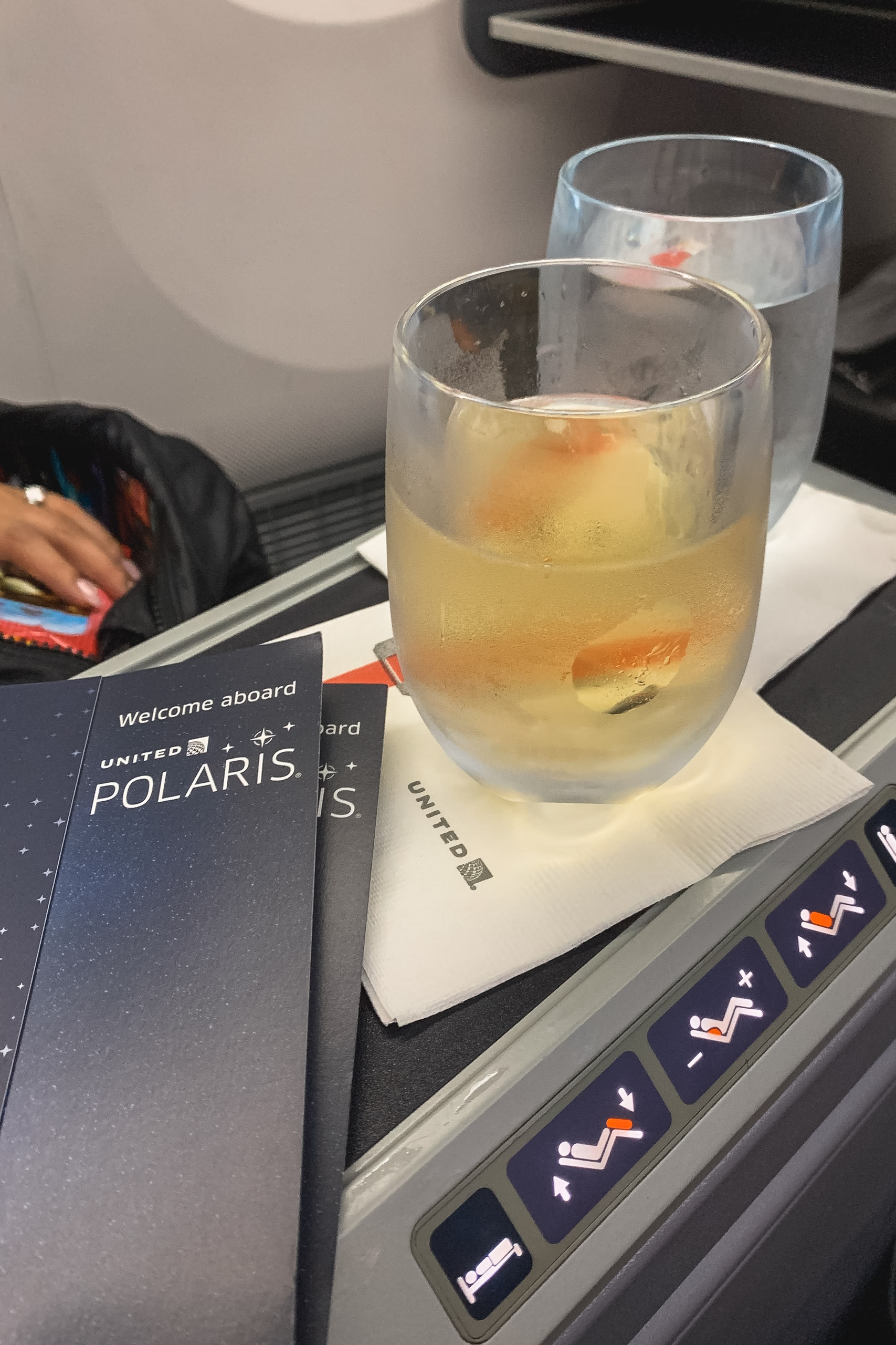 An airplane seat tray table holds a glass of amber-colored beverage with ice next to a United Airlines Polaris welcome card and empty glass during flight