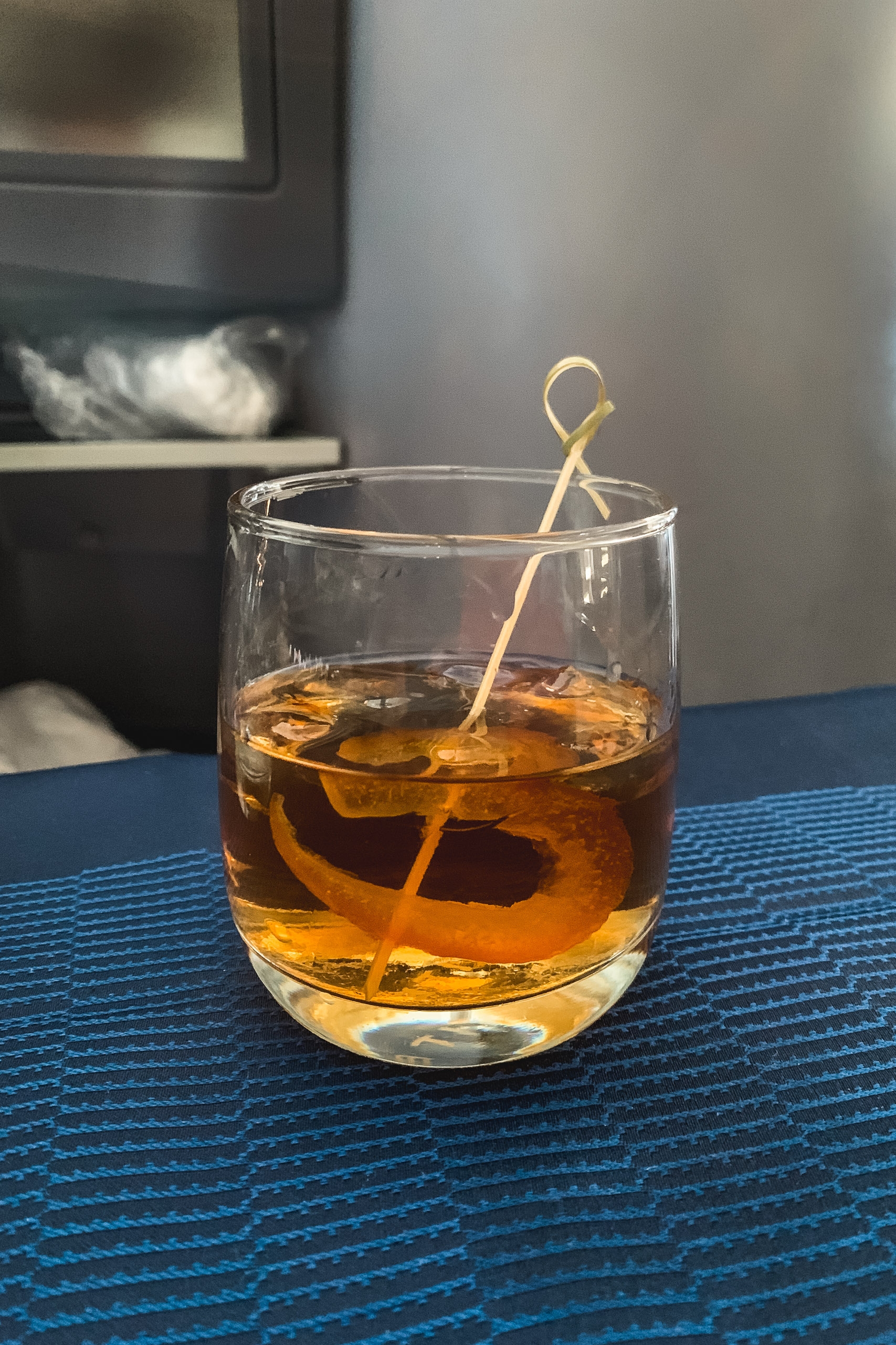 A glass of amber cocktail with ice and a twisted citrus peel stirrer sits on a blue textured surface, with blurred interior furnishings visible in the background